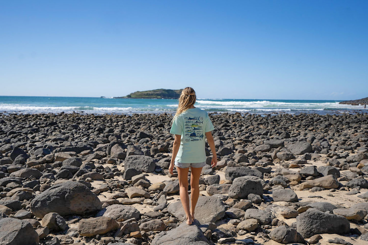 Person standing on a rocky beach in a Diver Mon Art top looking at the ocean under a clear blue sky.