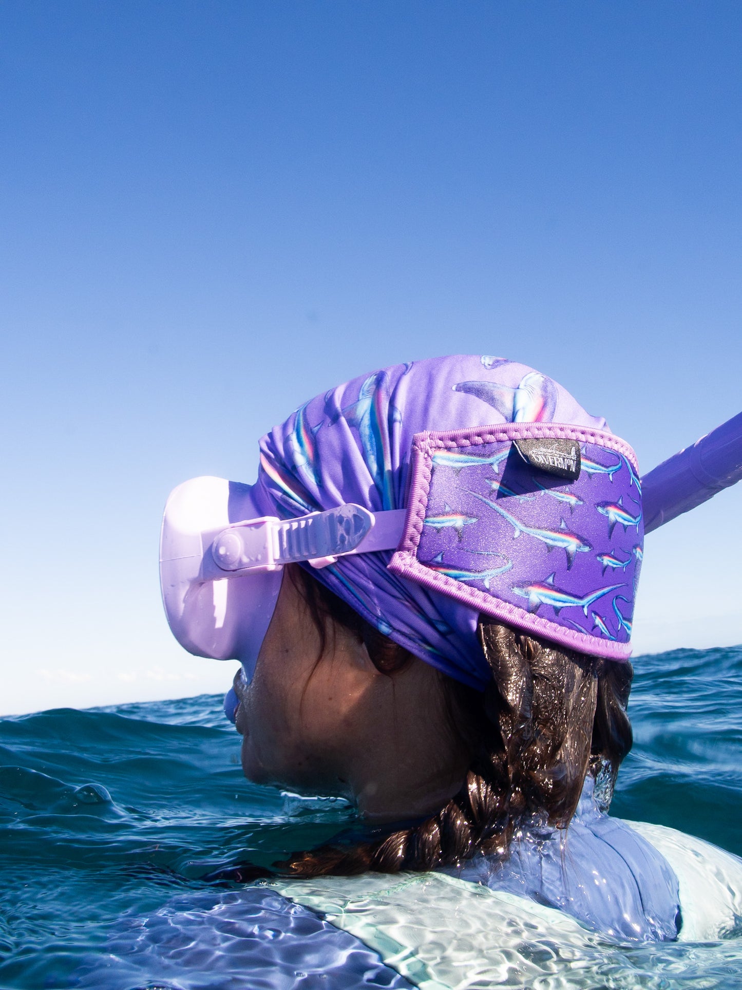 Dark haired woman seen close up side view in great detail above clear blue water wearing a purple mask fitted with the limited addition purple thresher neoprene mask tamer matching with the purple thresher UV gaiter ocean illustration by Diver Mon Art