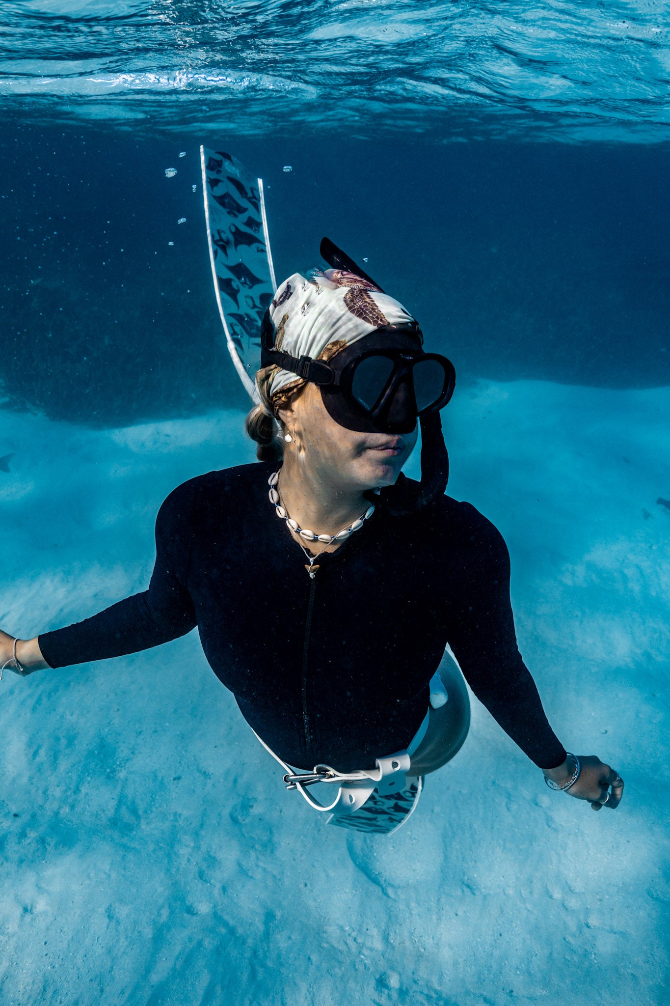 Blonde haired women shown underwater clear blue water with manta white fins wearing big turtles green UV gaiter buff design made from recycled polyester-spandex ocean illustration by Diver Mon Art