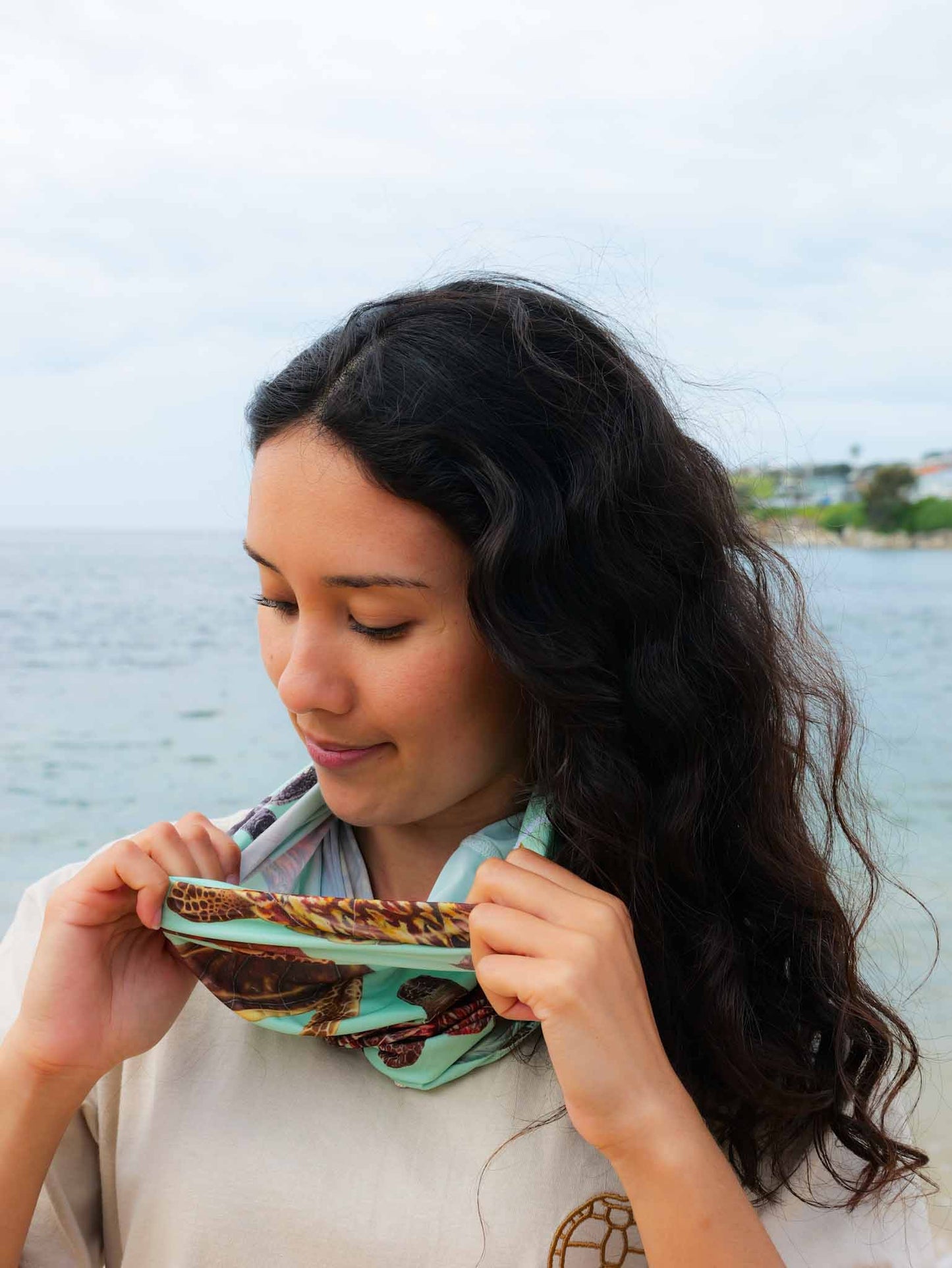 Dark haired women shown by at the beach putting on the big turtles green UV gaiter buff made from recycled polyester-spandex around bottom half of face neck ocean illustration by Diver Mon Art 