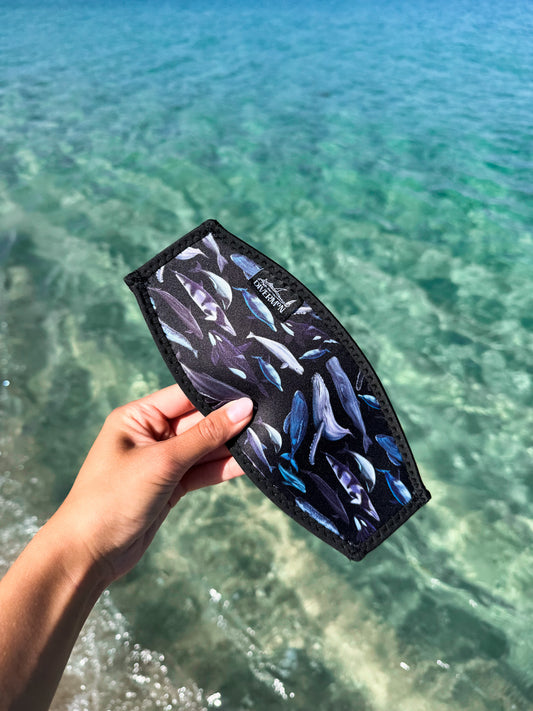 Hand holding a black neoprene mask tamer by Diver Mon Art with a diverse range of cetaceans species designs against a clear blue water background
