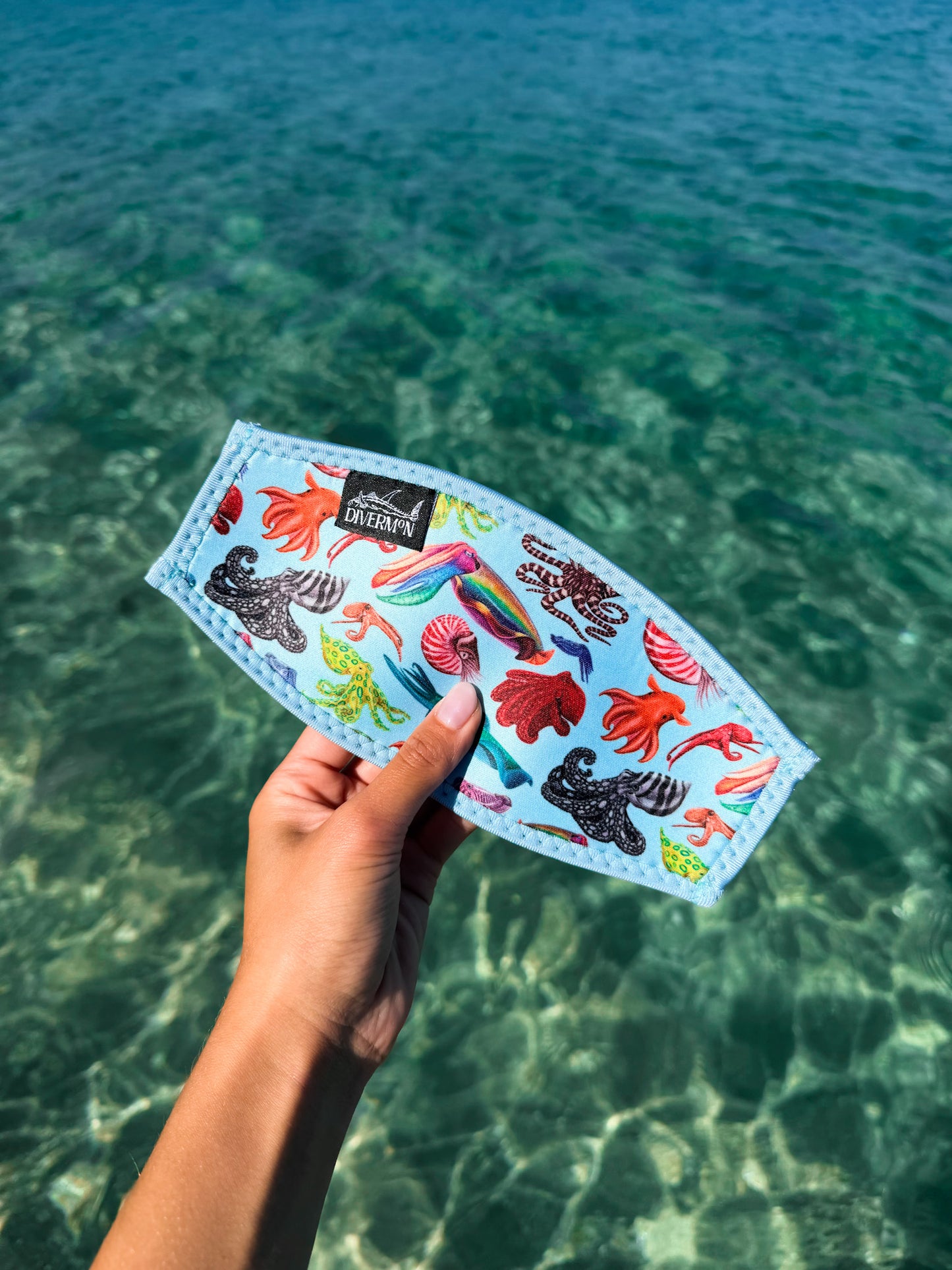 Hand holding a blue neoprene mask tamer by Diver Mon Art with a diverse range of cephalopod species designs against a clear blue water background