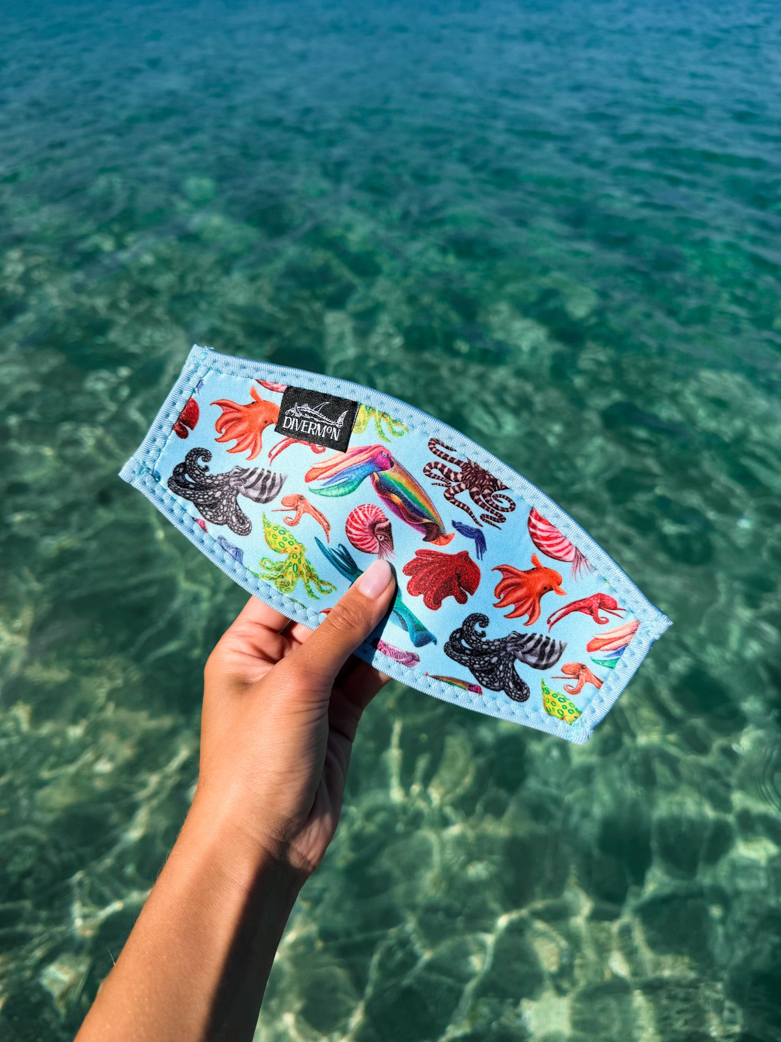 Hand holding a blue neoprene mask tamer by Diver Mon Art with a diverse range of cephalopod species designs against a clear blue water background
