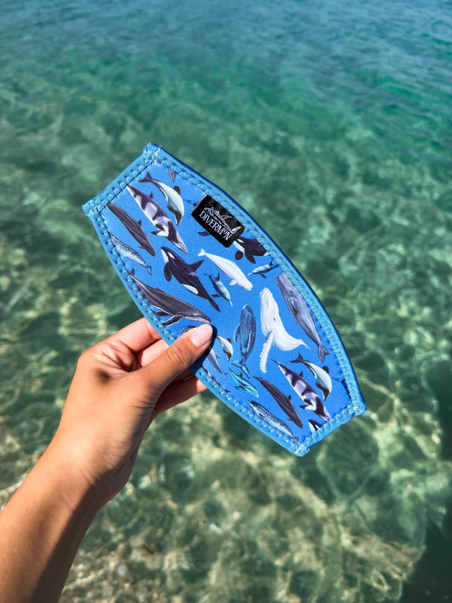 Hand holding a blue neoprene mask tamer by Diver Mon Art with a diverse range of cetaceans species designs against a clear blue water background