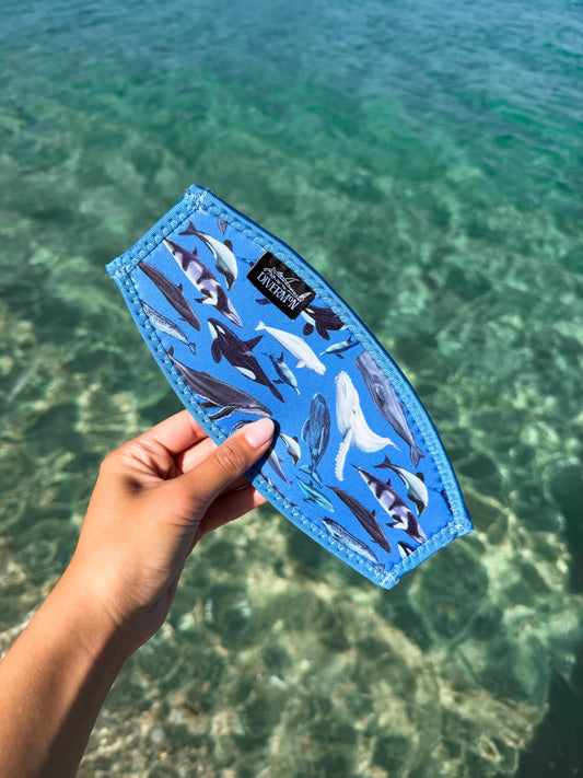 Hand holding a blue neoprene mask tamer by Diver Mon Art with a diverse range of cetaceans species designs against a clear blue water background