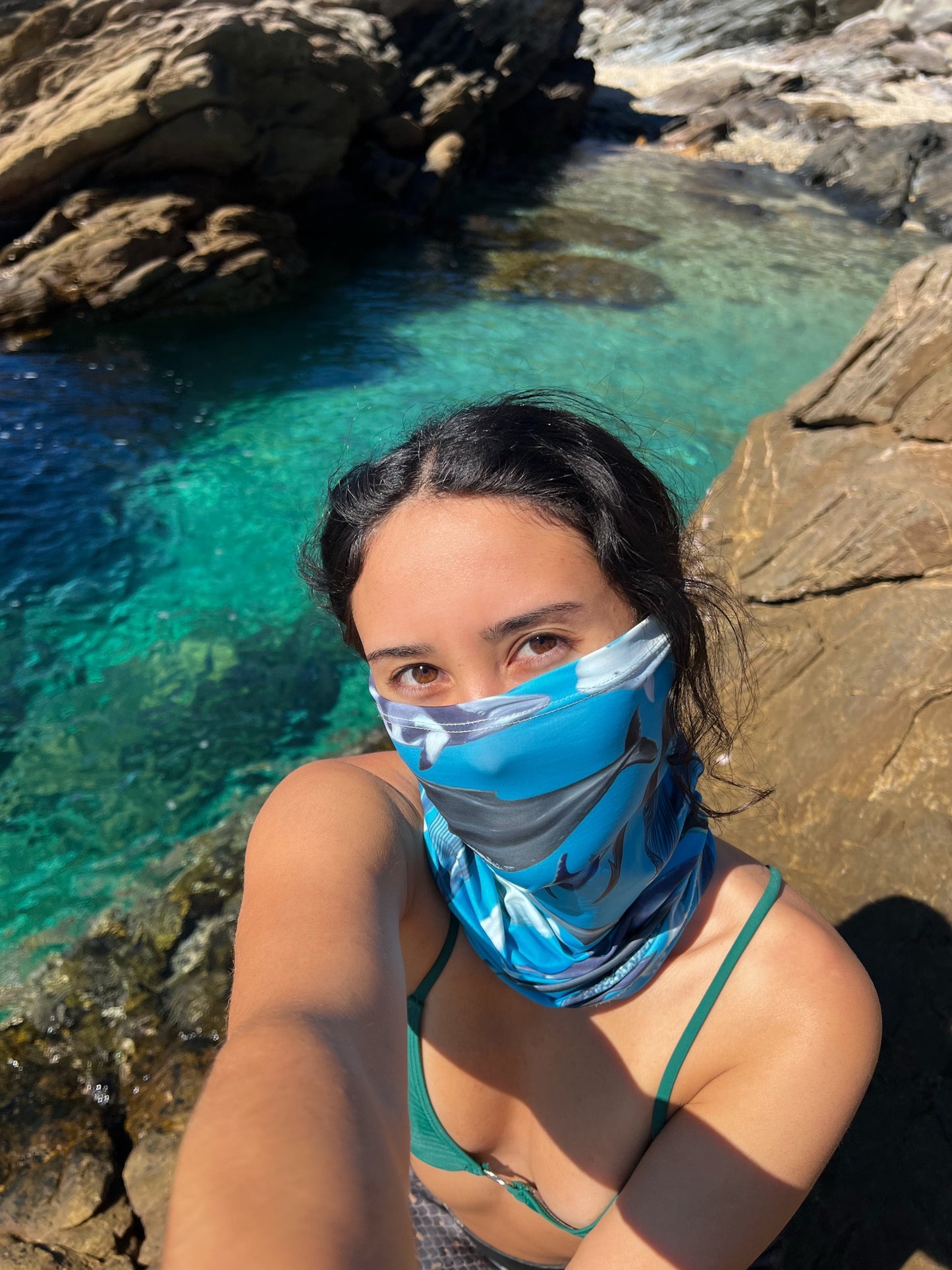 Dark haired women shown by a rock pool wearing the cetaceans blue UV gaiter buff made from recycled polyester-spandex around bottom half of face neck ocean illustration by Diver Mon Art 