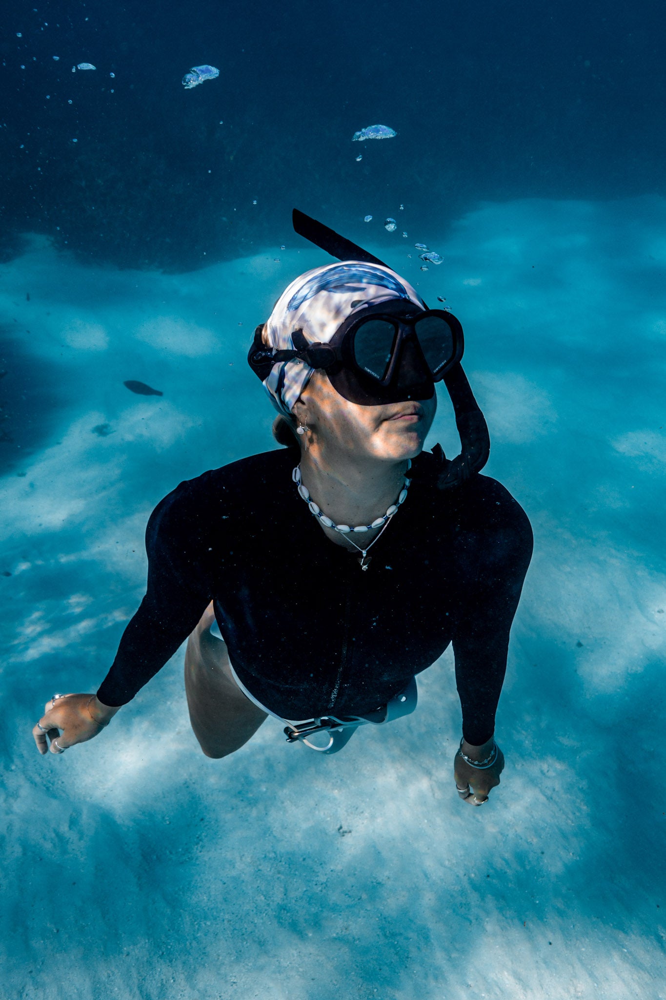 Blonde haired women shown underwater clear blue water wearing cetaceans white UV gaiter buff design made from recycled polyester-spandex ocean illustration by Diver Mon Art