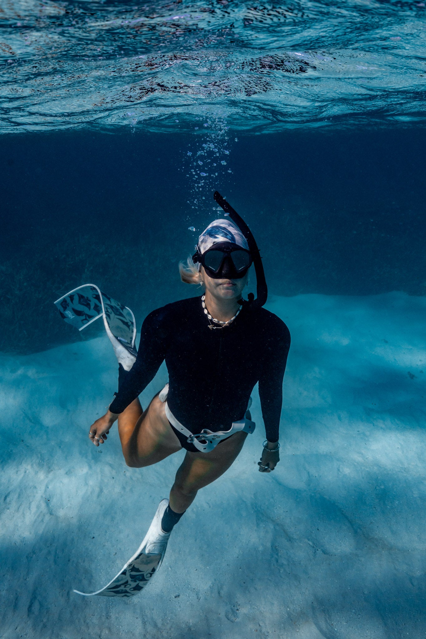 Blonde haired women shown from afar underwater clear blue water with manta white fins wearing cetaceans white UV gaiter buff design made from recycled polyester-spandex ocean illustration by Diver Mon Art