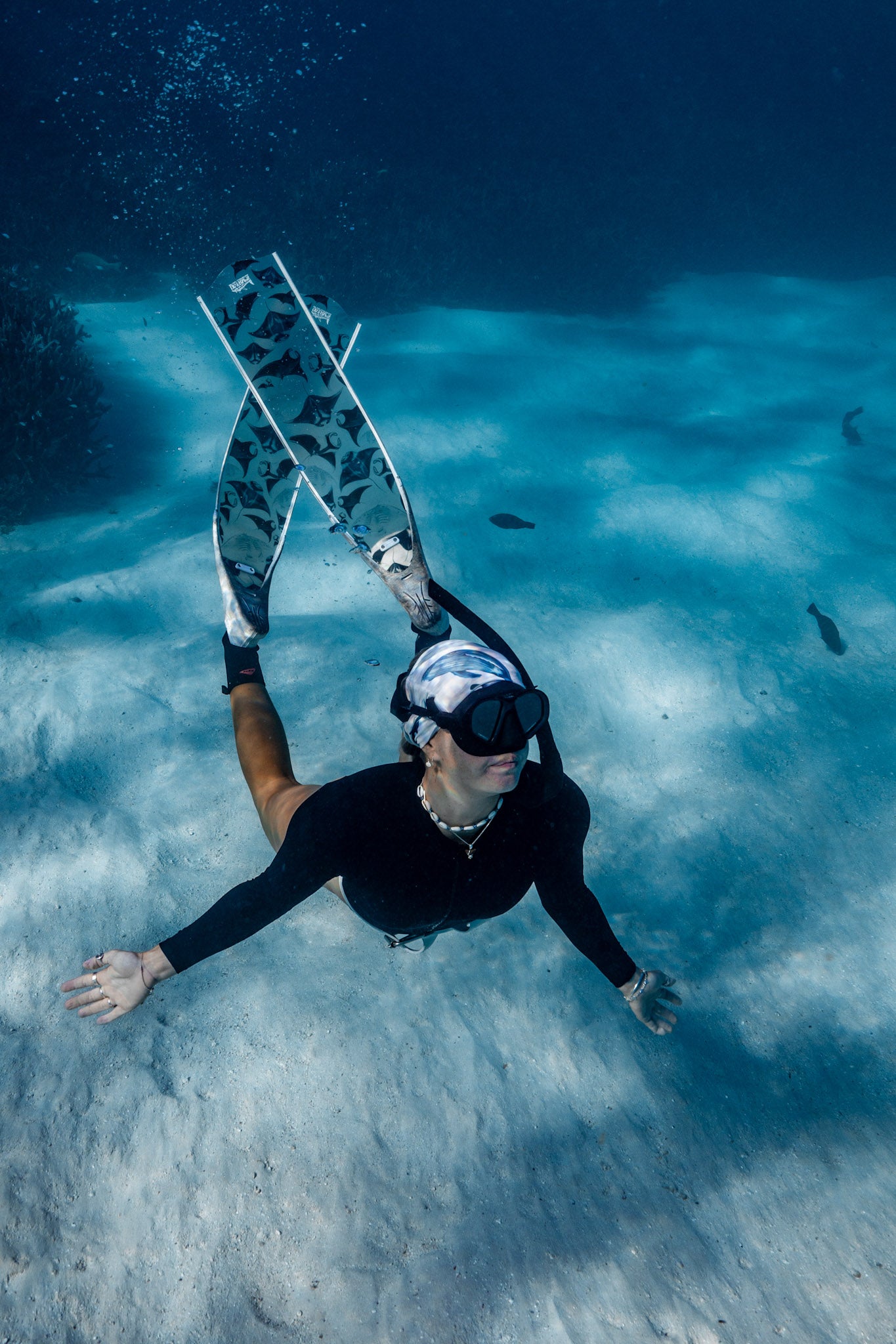 Blonde haired women shown from afar full body underwater clear blue water with manta white fins wearing cetaceans white UV gaiter buff design made from recycled polyester-spandex ocean illustration by Diver Mon Art