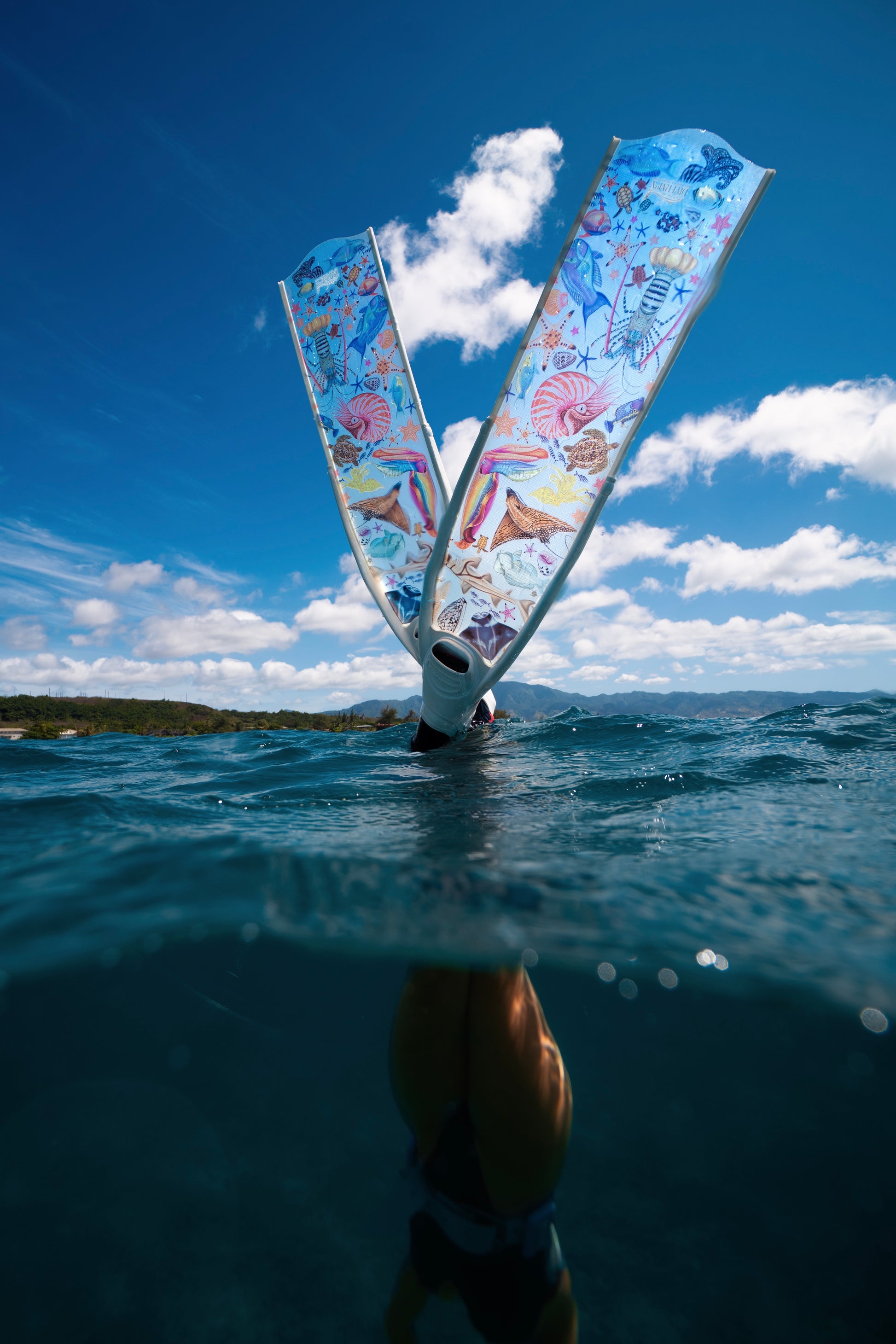 Person underwater with a colorful freediving fins in the air against a blue sky with clouds.