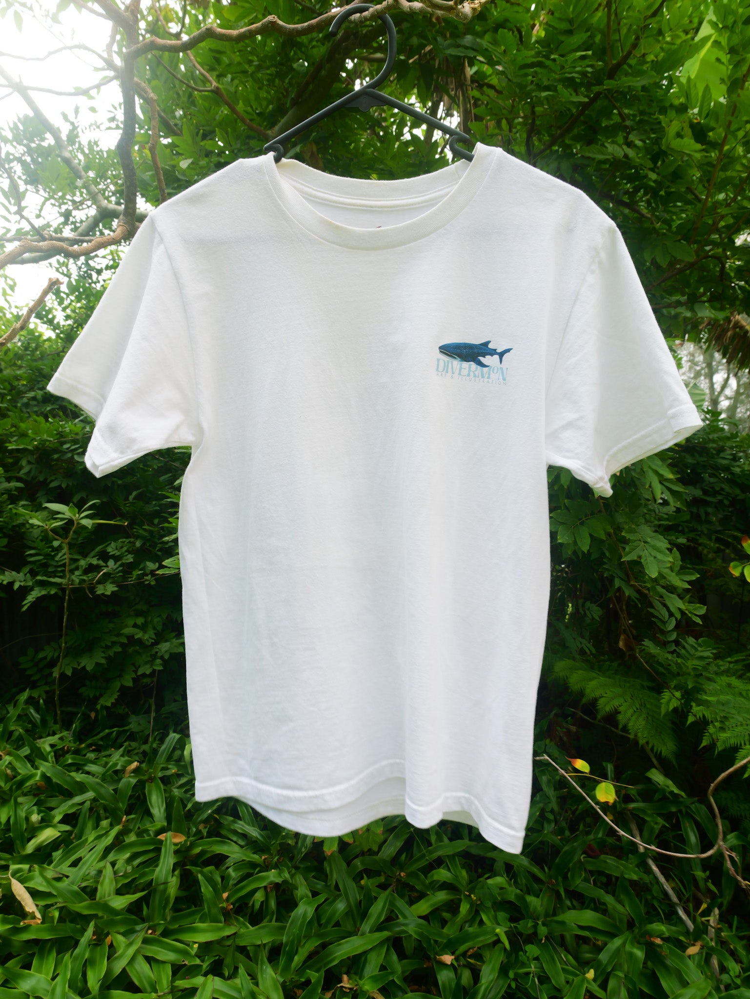 White t-shirt with a blue whaleshark graphic on a hanger against a green leafy background