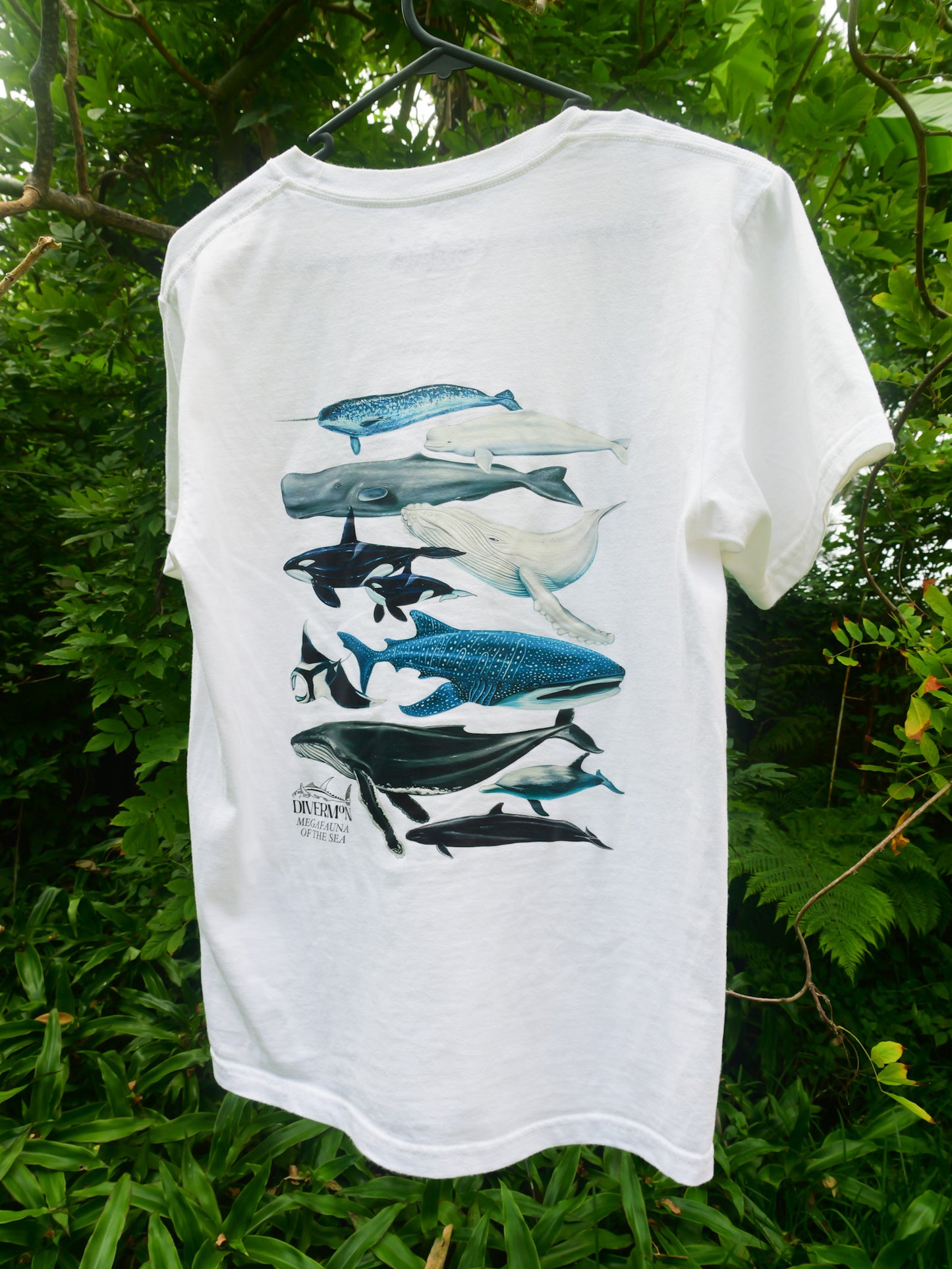 White t-shirt with whale and fish illustrations on a hanger against a green leafy background