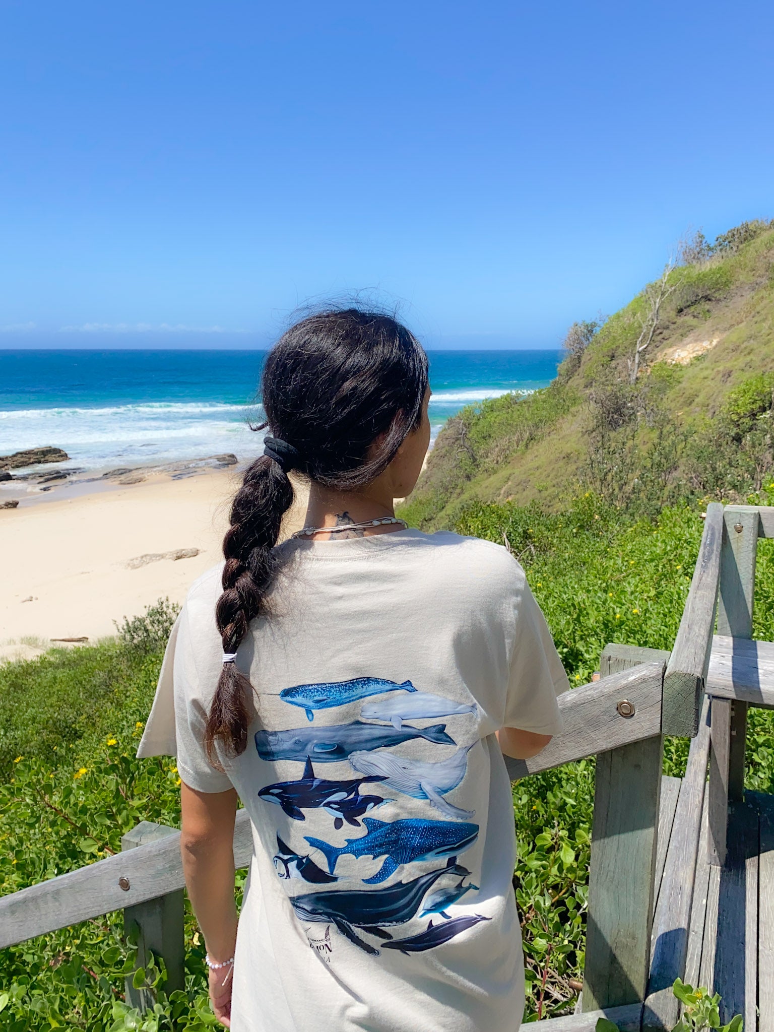 Person wearing a tan t-shirt with blue whale designs, standing on a beach with ocean and greenery in the background.