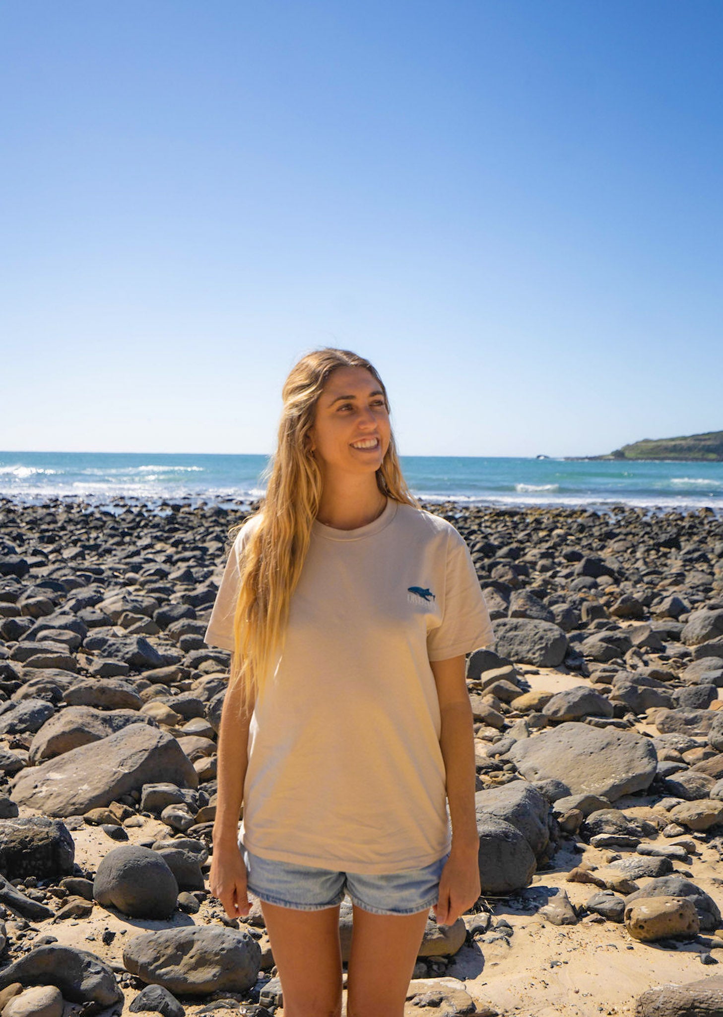 Woman standing on a rocky beach wearing a Diver Mon Art tee with ocean and clear blue sky in the background