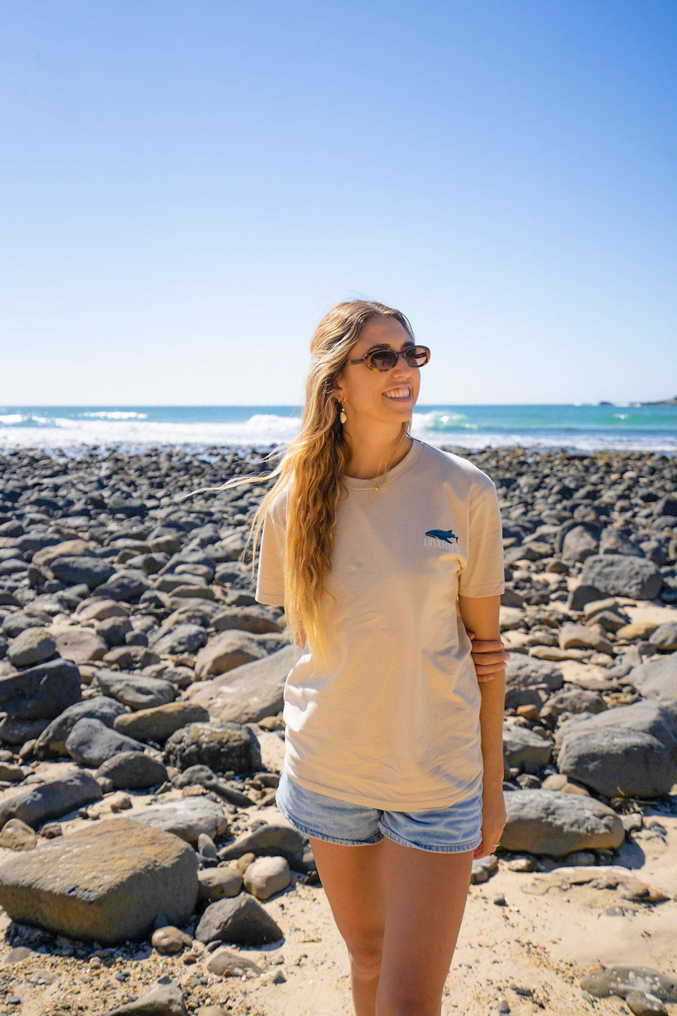 Woman standing on a rocky beach wearing sunglasses and a tan-colored t-shirt. Diver Mon Art
