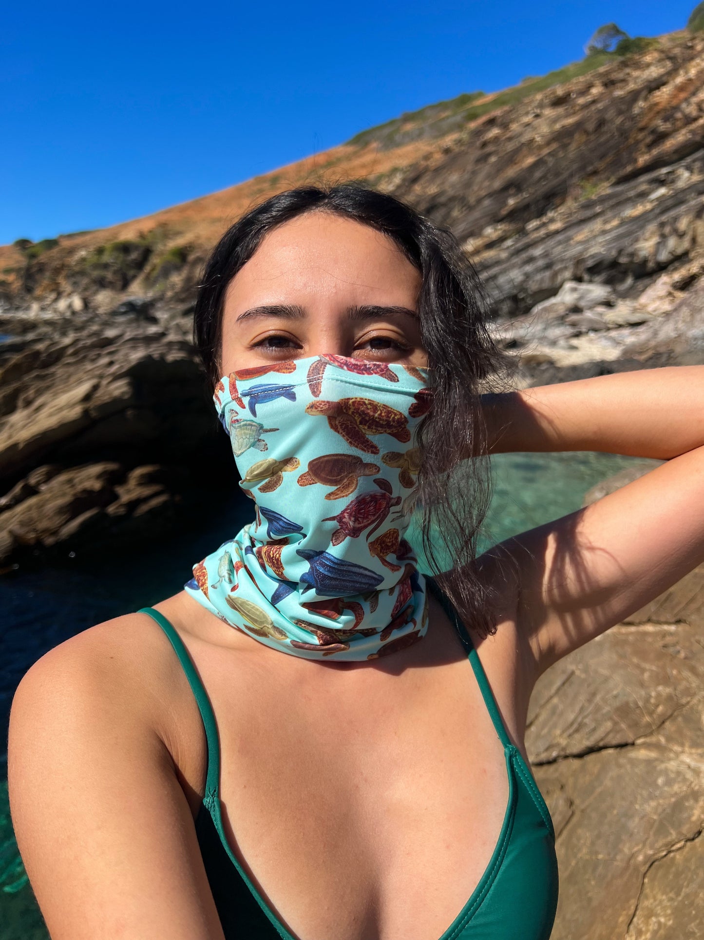 Dark haired women shown by a rock pool wearing the mini turtles green UV gaiter buff made from recycled polyester-spandex around bottom half of face neck ocean illustration by Diver Mon Art