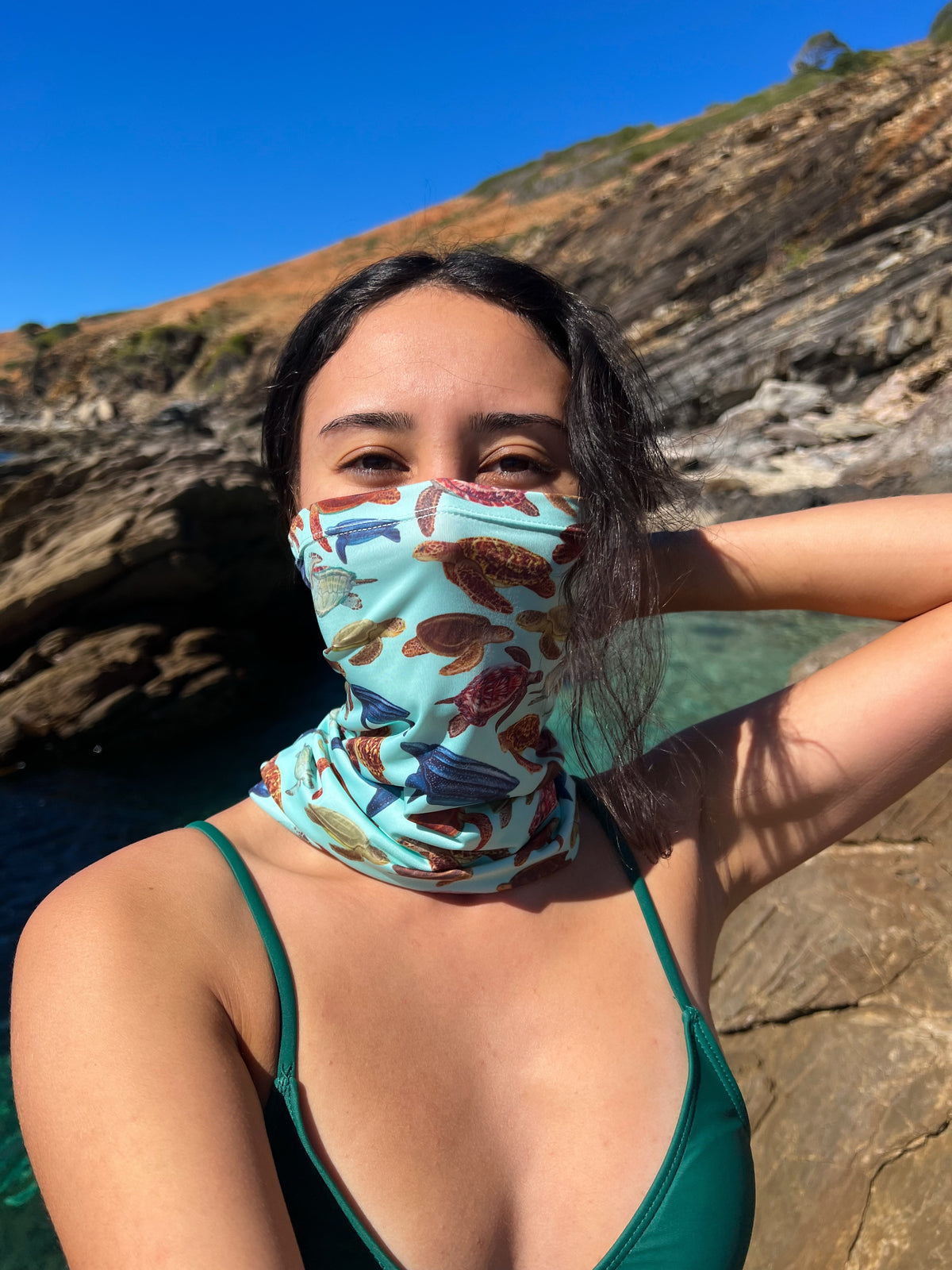 Dark haired women shown by a rock pool wearing the mini turtles green UV gaiter buff made from recycled polyester-spandex around bottom half of face neck ocean illustration by Diver Mon Art