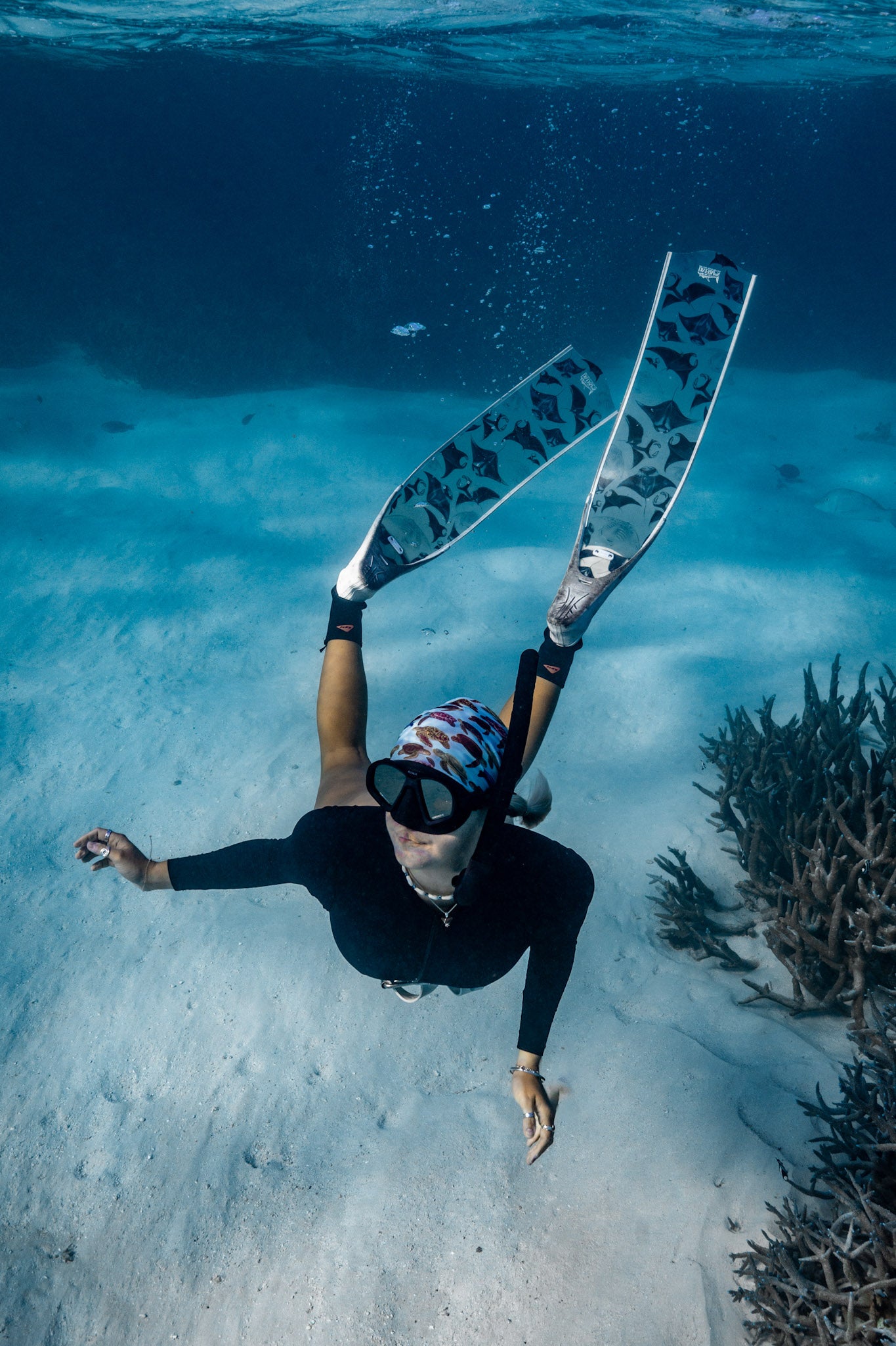 Blonde haired women shown from afar underwater clear blue water with manta white fins wearing mini turtles green UV gaiter buff design made from recycled polyester-spandex ocean illustration by Diver Mon Art