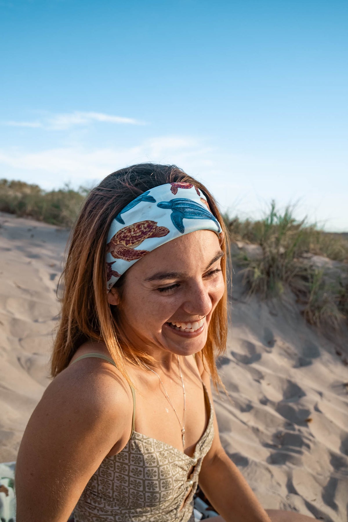 Dark haired women smiling shown close up at the beach in the sunlight wearing the mini turtles green UV gaiter buff design made from recycled polyester-spandex  in a headband way ocean illustration made by Diver Mon Art