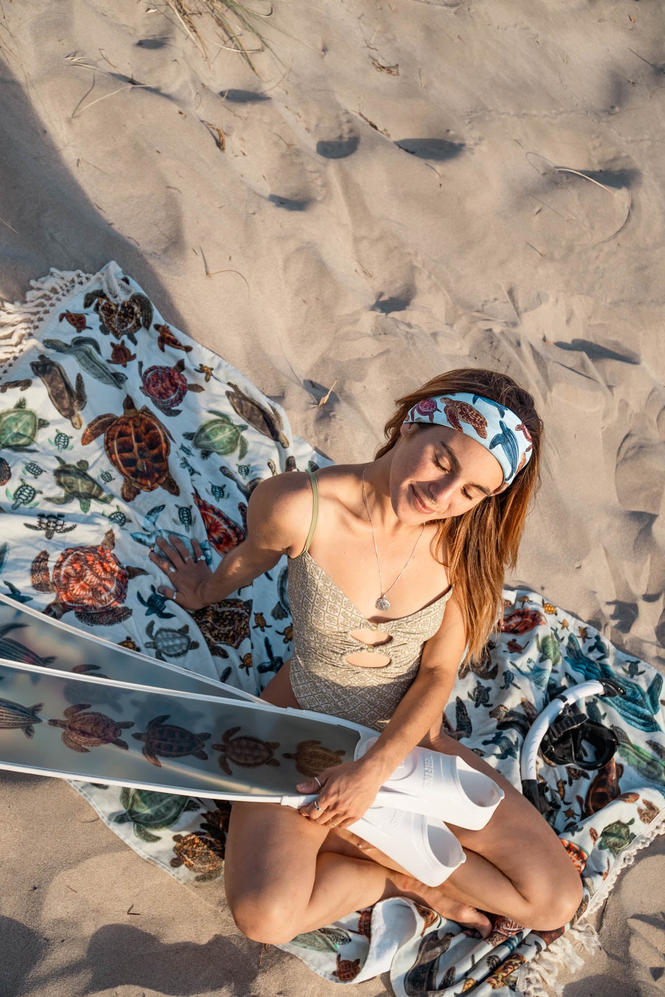 Dark haired women shown at the beach in the sunlight sitting on XL turtle towel, wearing the mini turtles green UV gaiter buff design made from recycled polyester-spandex in a headband way while holding Diver Mon turtle freediving fins with white rails ocean illustration made by Diver Mon Art