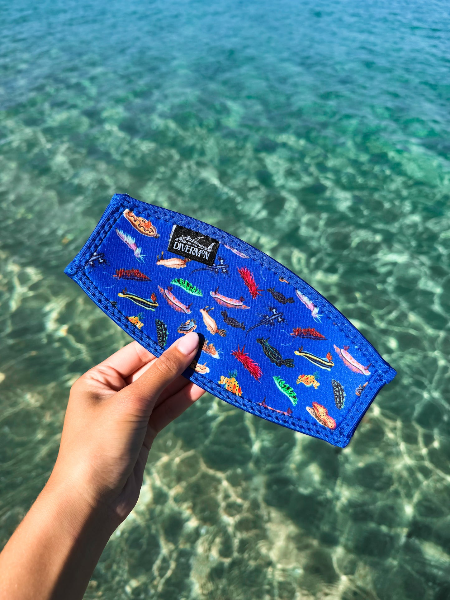 Hand holding a dark blue nudibranch neoprene mask tamer by Diver Mon Art design against a clear blue water background