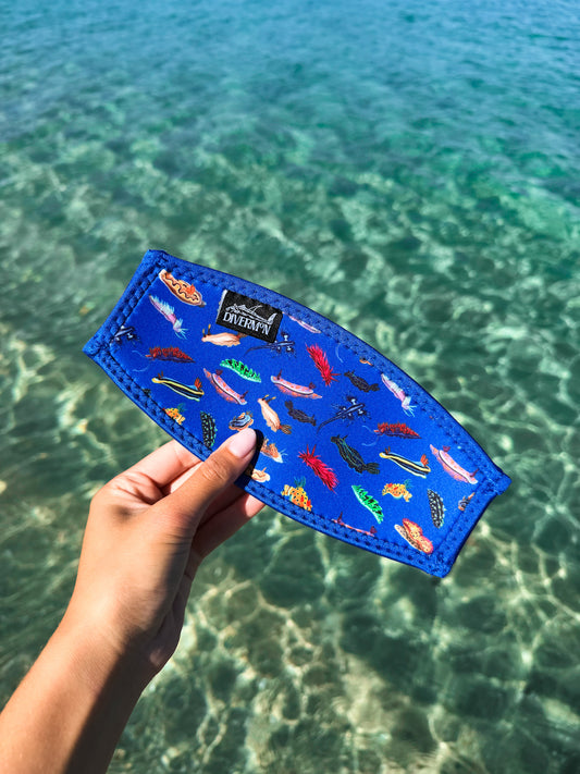 Hand holding a dark blue nudibranch neoprene mask tamer by Diver Mon Art design against a clear blue water background