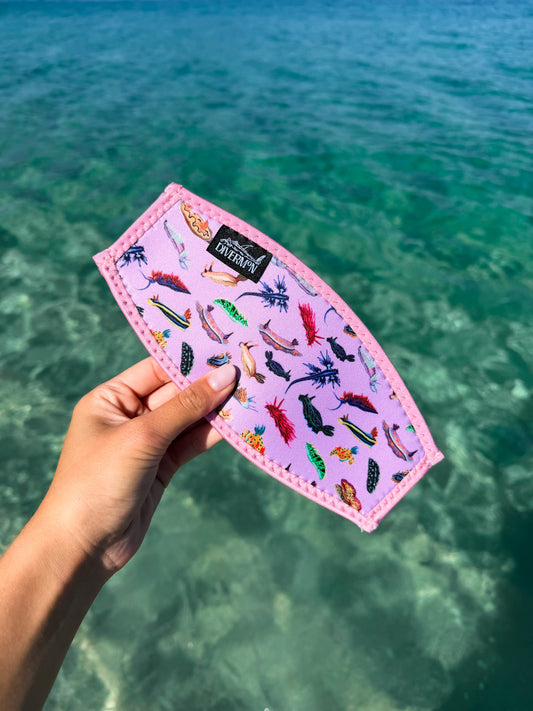 Hand holding nudibranch pink neoprene mask tamer design by Diver Mon Art design against a clear blue water background