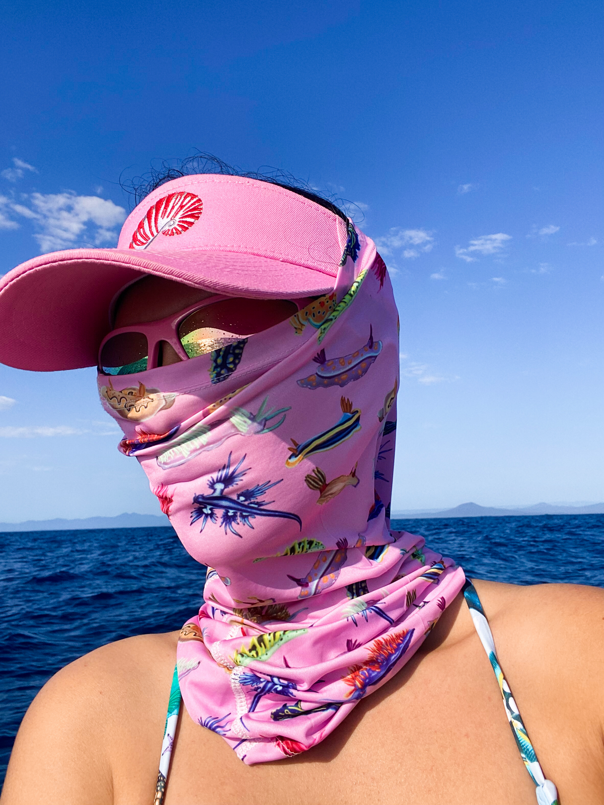 Dark haired women shown on the ocean wearing the nudibranch pink UV gaiter buff made from recycled polyester-spandex around bottom half of face neck along with nautilus visor hat ocean illustration by Diver Mon Art