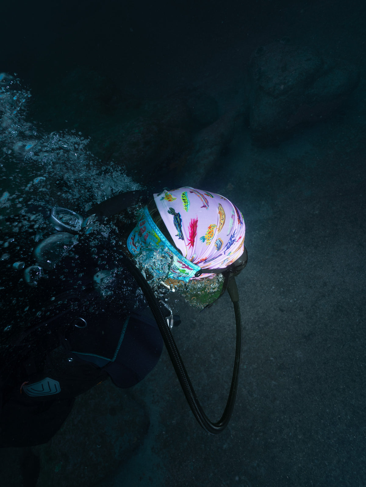 Scuba diver shown close up from behind underwater wearing nudibranch pink UV gaiter buff design made from recycled polyester-spandex along with the seahorse green tamer made from neoprene ocean illustration by Diver Mon Art