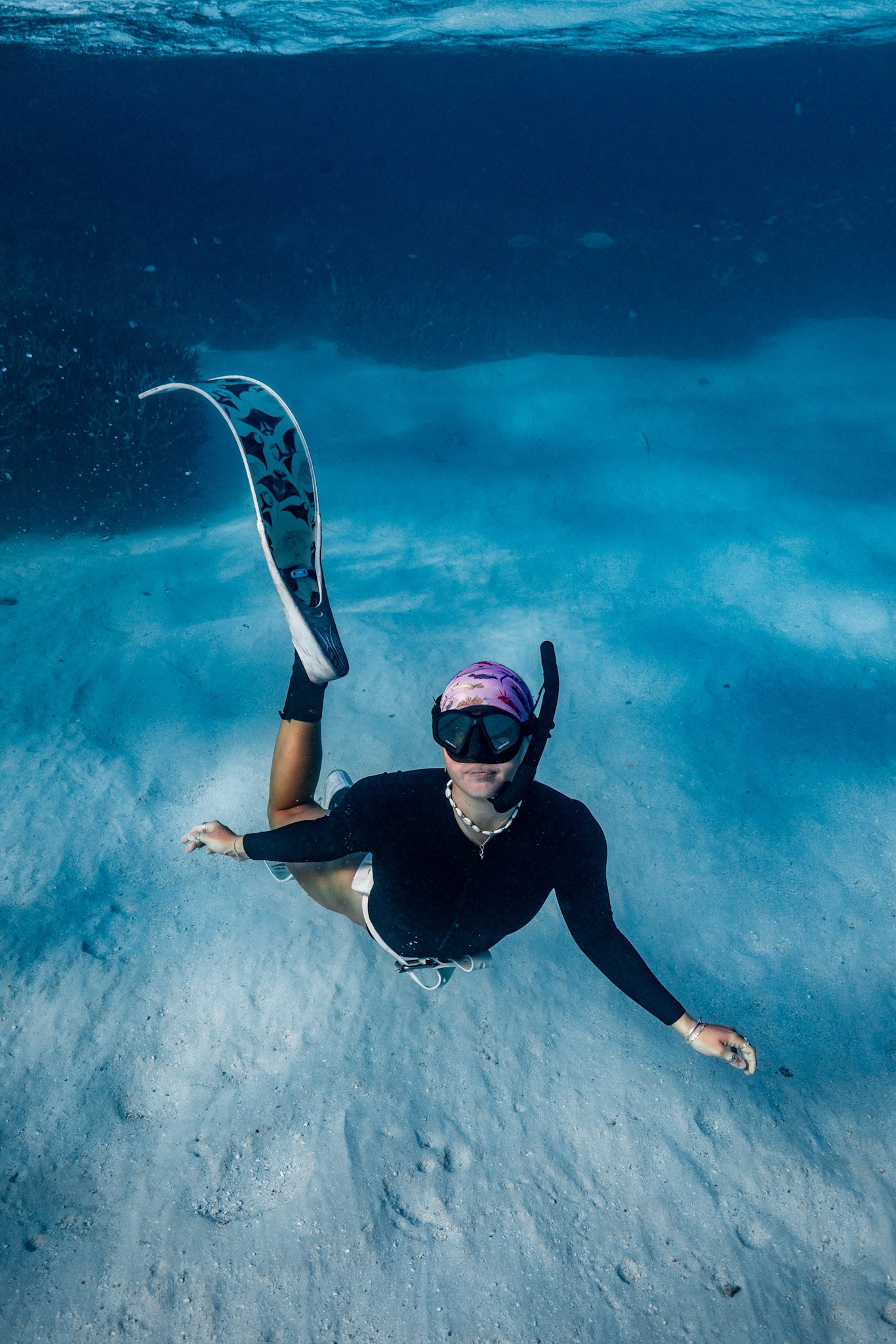 Blonde haired women shown from afar underwater clear blue water with manta white fins wearing nudibranch pink UV gaiter buff design made from recycled polyester-spandex ocean illustration by Diver Mon Art