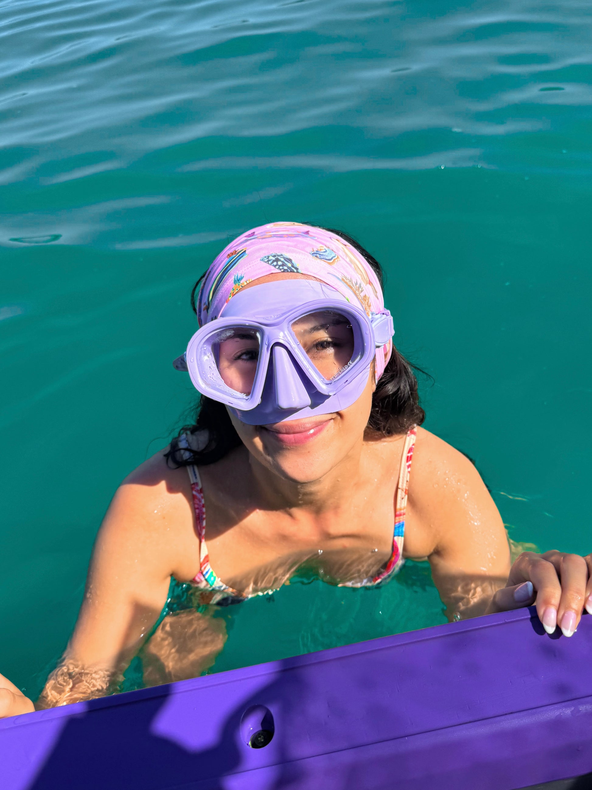 Dark haired women smiling shown close up in the water and sunlight with purple mask on wearing the nudibranch pink UV gaiter buff design made from recycled polyester-spandex  in a headband way ocean illustration made by Diver Mon Art