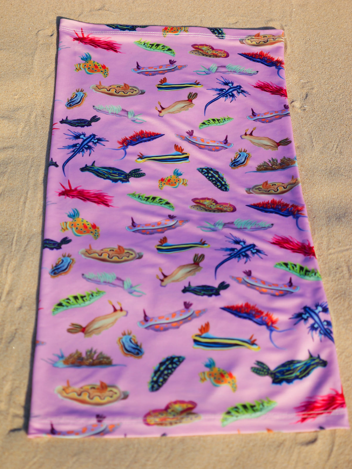 Nudibranch pink UV gaiter buff design shown laid out full length close up in great detail sand background made from recycled polyester-spandex ocean illustration by Diver Mon Art