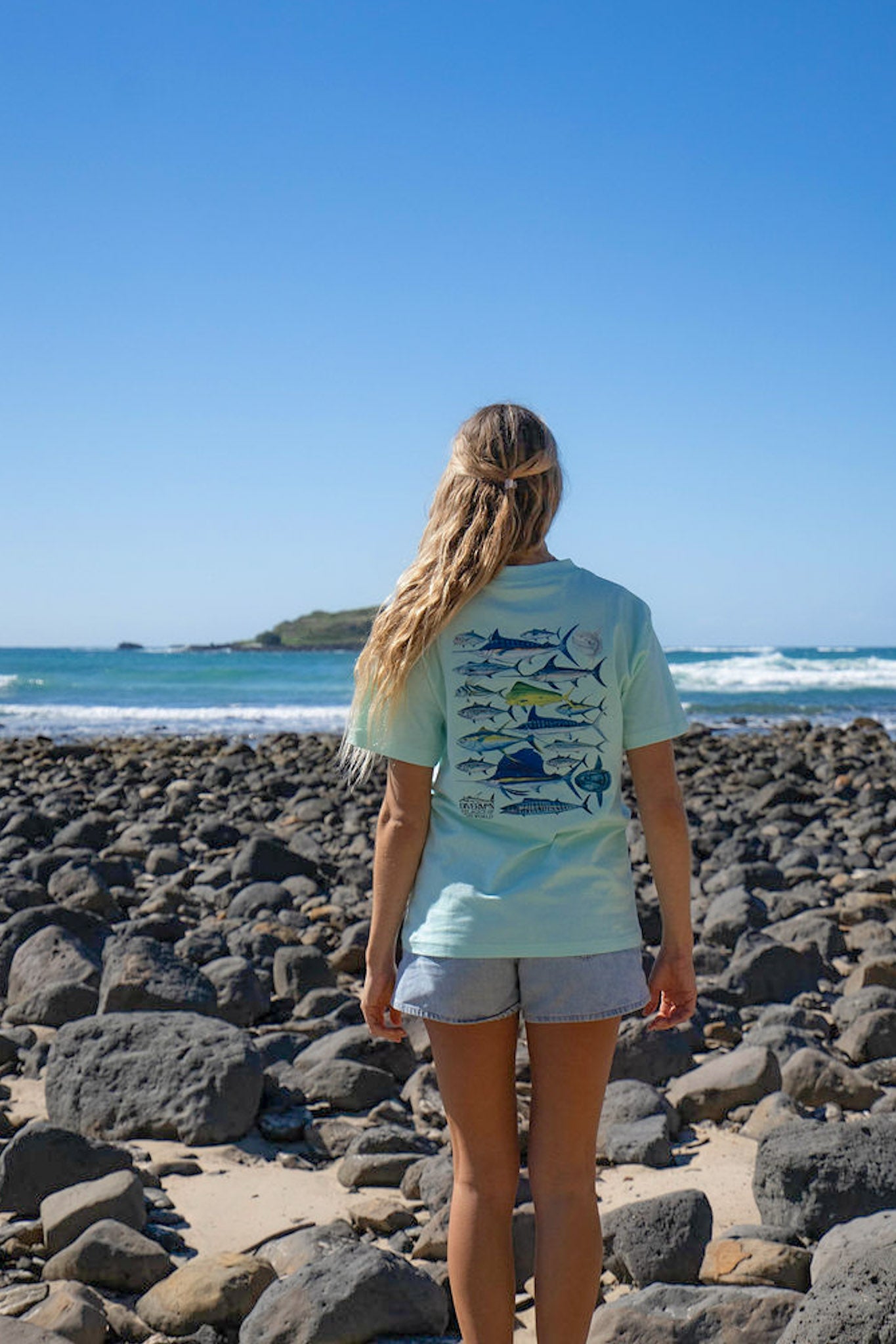 Person wearing a light blue t-shirt with pelagic fish designs on a rocky beach.