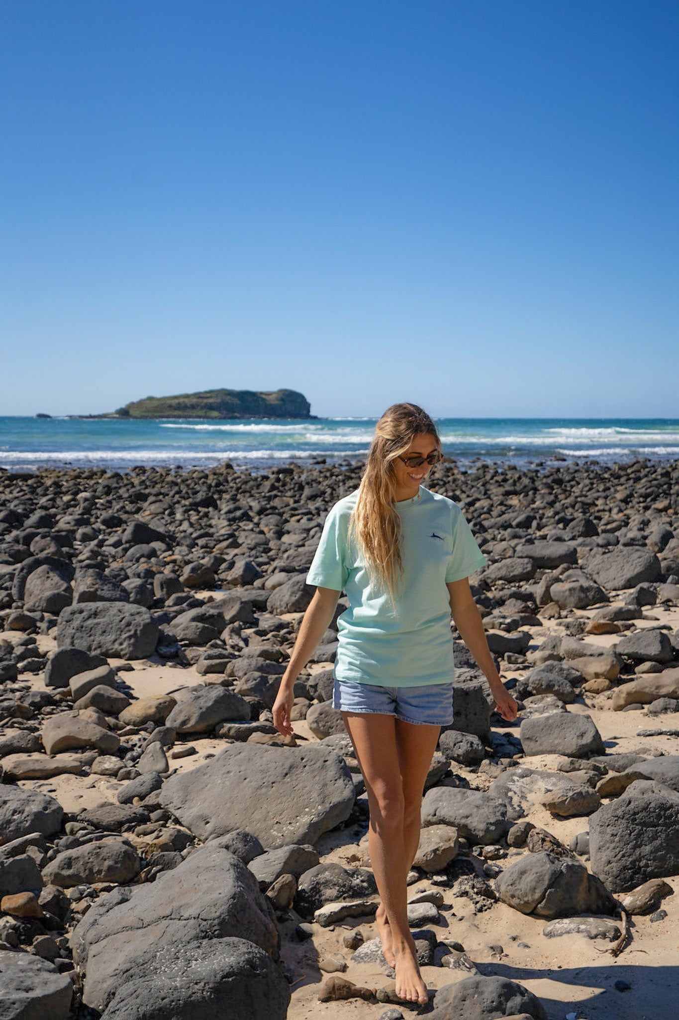 Woman walking on a rocky beach wearing a Diver Mon Art top clear blue sky and ocean in the background