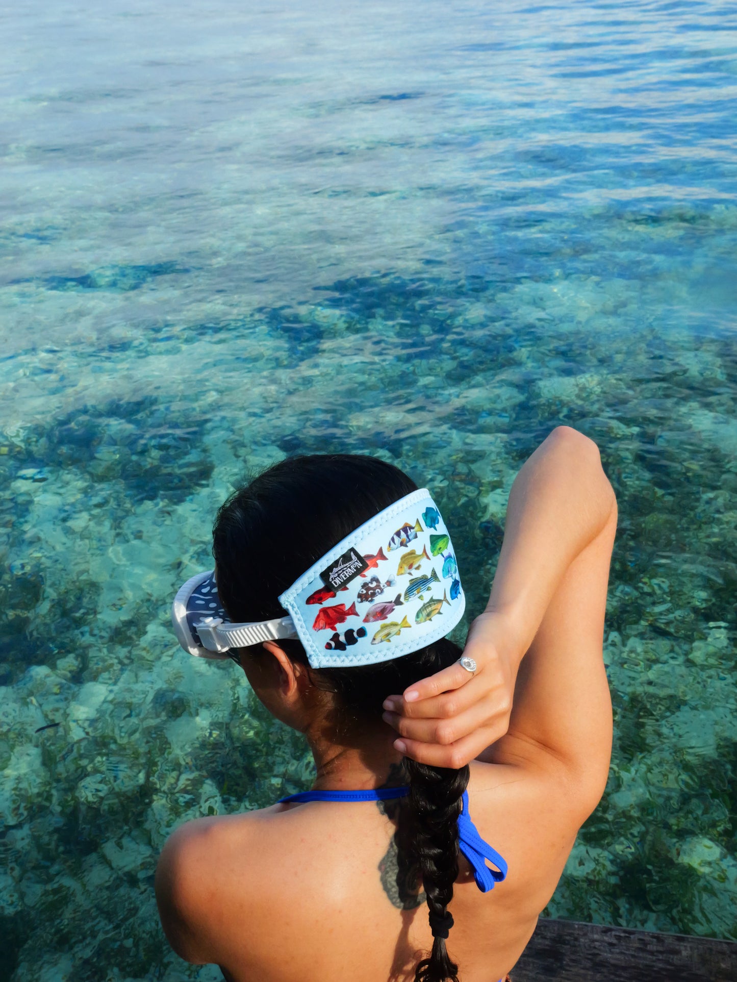 Dark haired woman seen from behind in clear blue water wearing a mask fitted with the rainbow fish design with blue background neoprene mask strap tamer ocean illustration by Diver Mon Art 