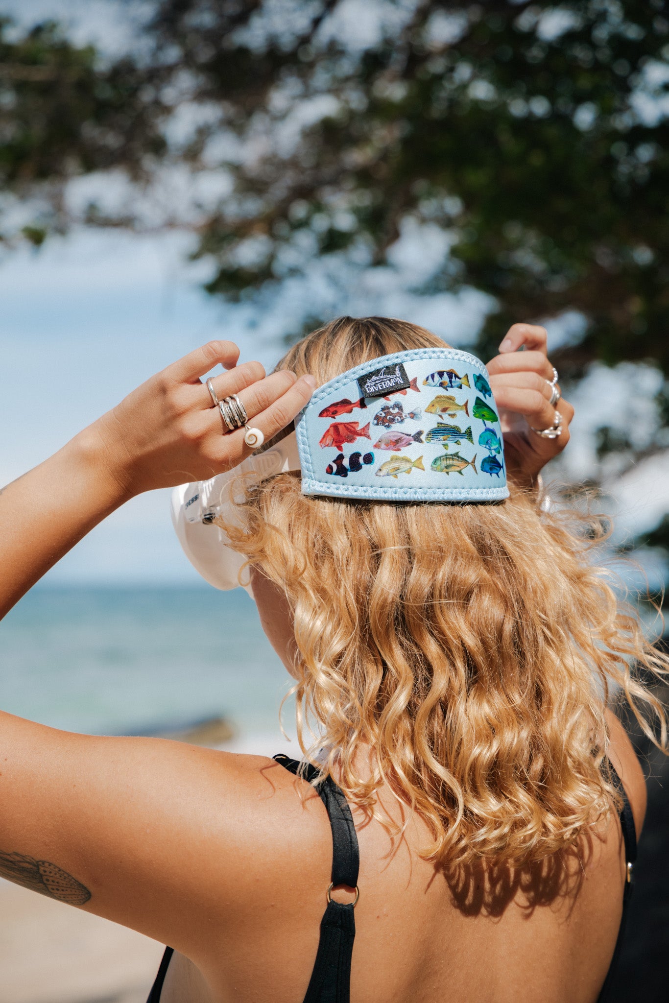 Blonde haired woman seen close up side view by the beach with the rainbow fish blue design neoprene mask strap tamer fitted to the mask ocean illustration by Diver Mon Art 