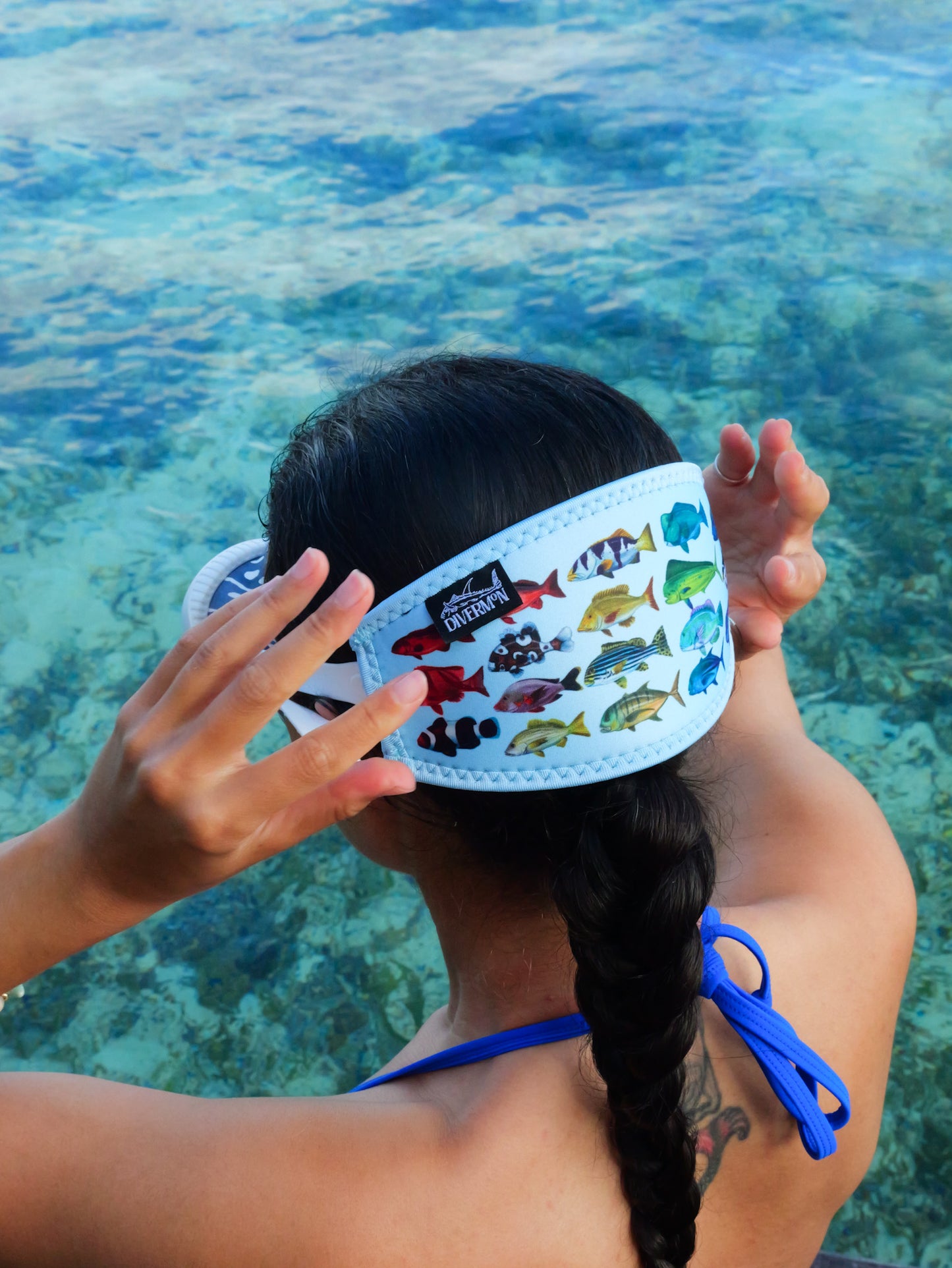 Dark haired woman shown from behind in blue clear water close up wearing a mask fitted with the rainbow fish blue design neoprene mask strap tamer ocean illustration by Diver Mon Art