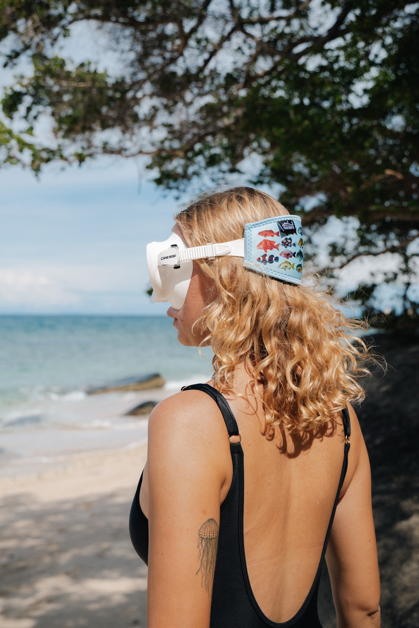 Blonde haired woman seen from the side at the beach wearing the rainbow fish blue design neoprene mask strap tamer fitted to mask ocean illustration by Diver Mon Art