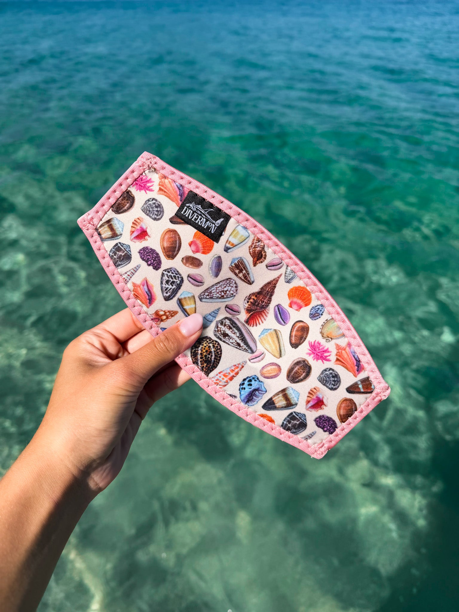Hand holding a pink neoprene mask tamer by Diver Mon Art with colourful shell designs against a clear blue water background