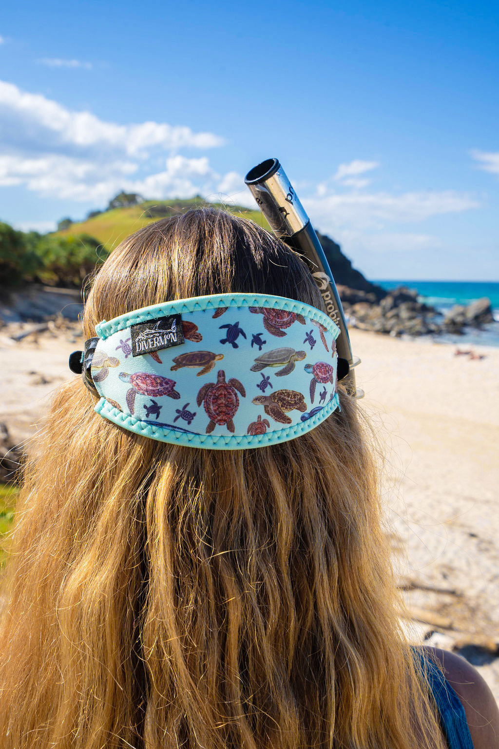 Blonde haired woman seen close up from behind at the beach with the green turtle design neoprene mask strap tamer ocean illustration by Diver Mon Art 