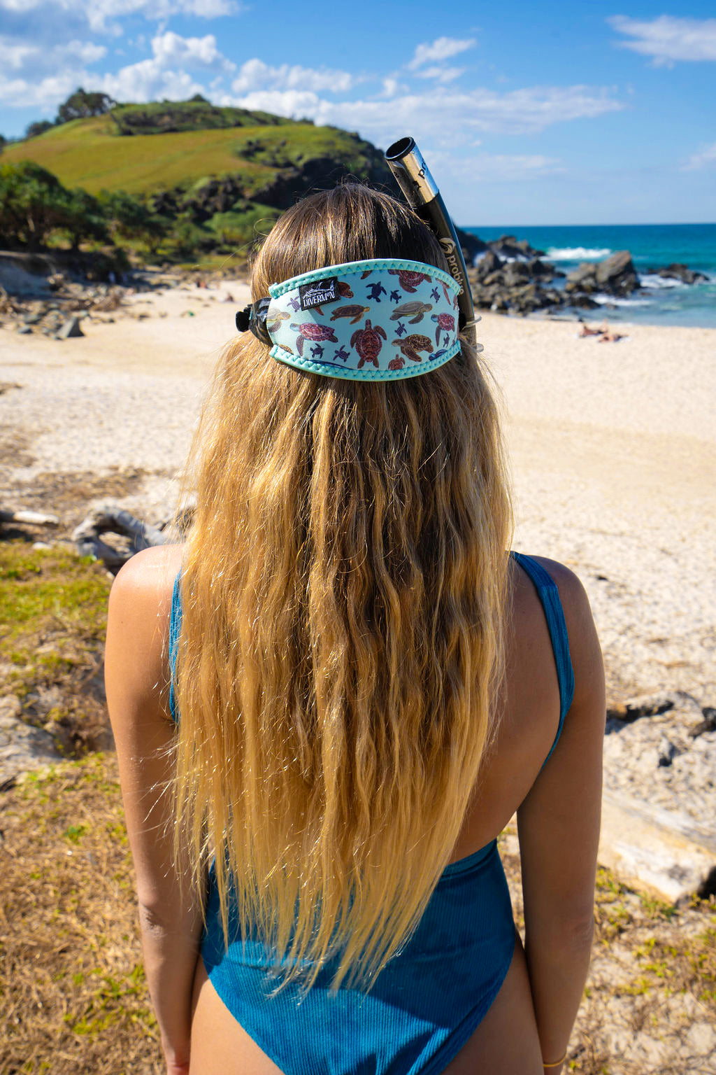 Blonde haired woman seen close up from behind at the beach with the green turtle design neoprene mask strap tamer ocean illustration by Diver Mon Art 