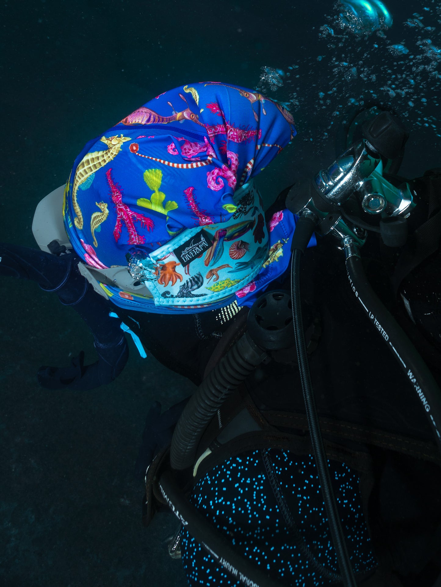 Scuba diver shown close up underwater wearing seahorse dark blue UV gaiter buff design made from recycled polyester-spandex along with the cephalopod blue tamer made from neoprene ocean illustration by Diver Mon Art