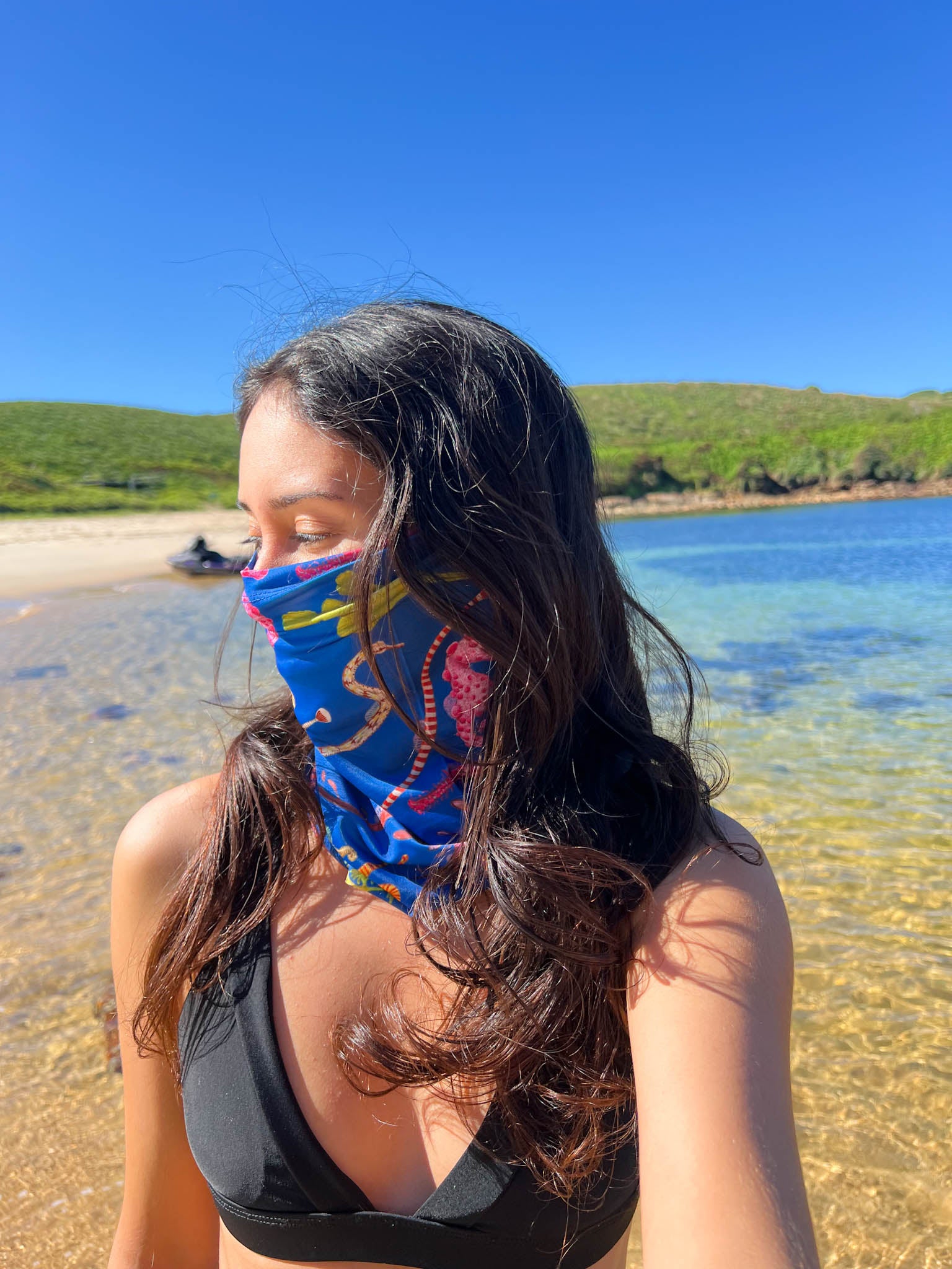 Dark haired women shown by the beach shoreline wearing the seahorse dark blue UV gaiter buff made from recycled polyester-spandex around bottom half of face neck ocean illustration by Diver Mon Art