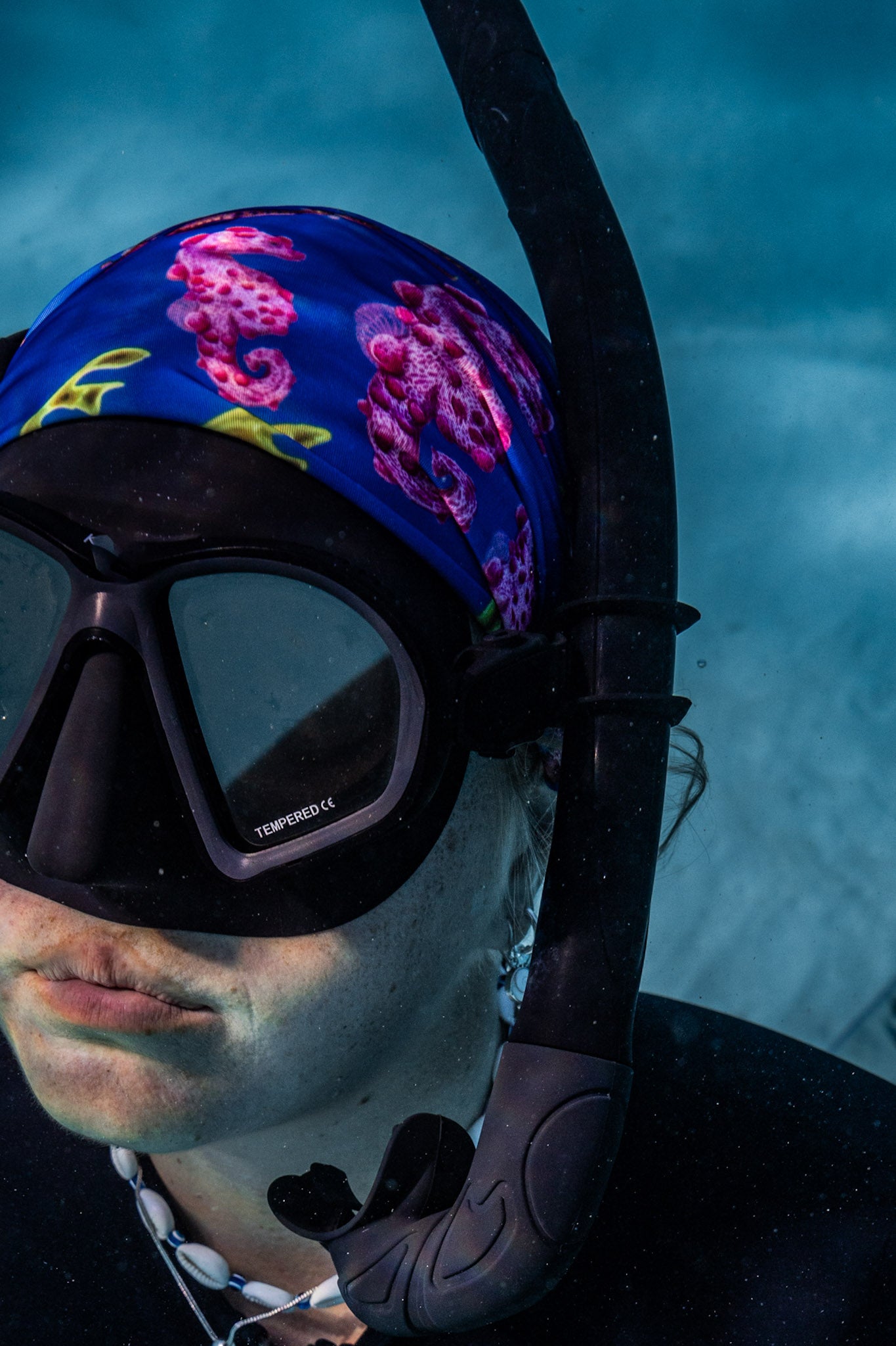 Blonde haired women shown close up great detail underwater clear blue water wearing seahorse dark blue UV gaiter buff design made from recycled polyester-spandex ocean illustration by Diver Mon Art