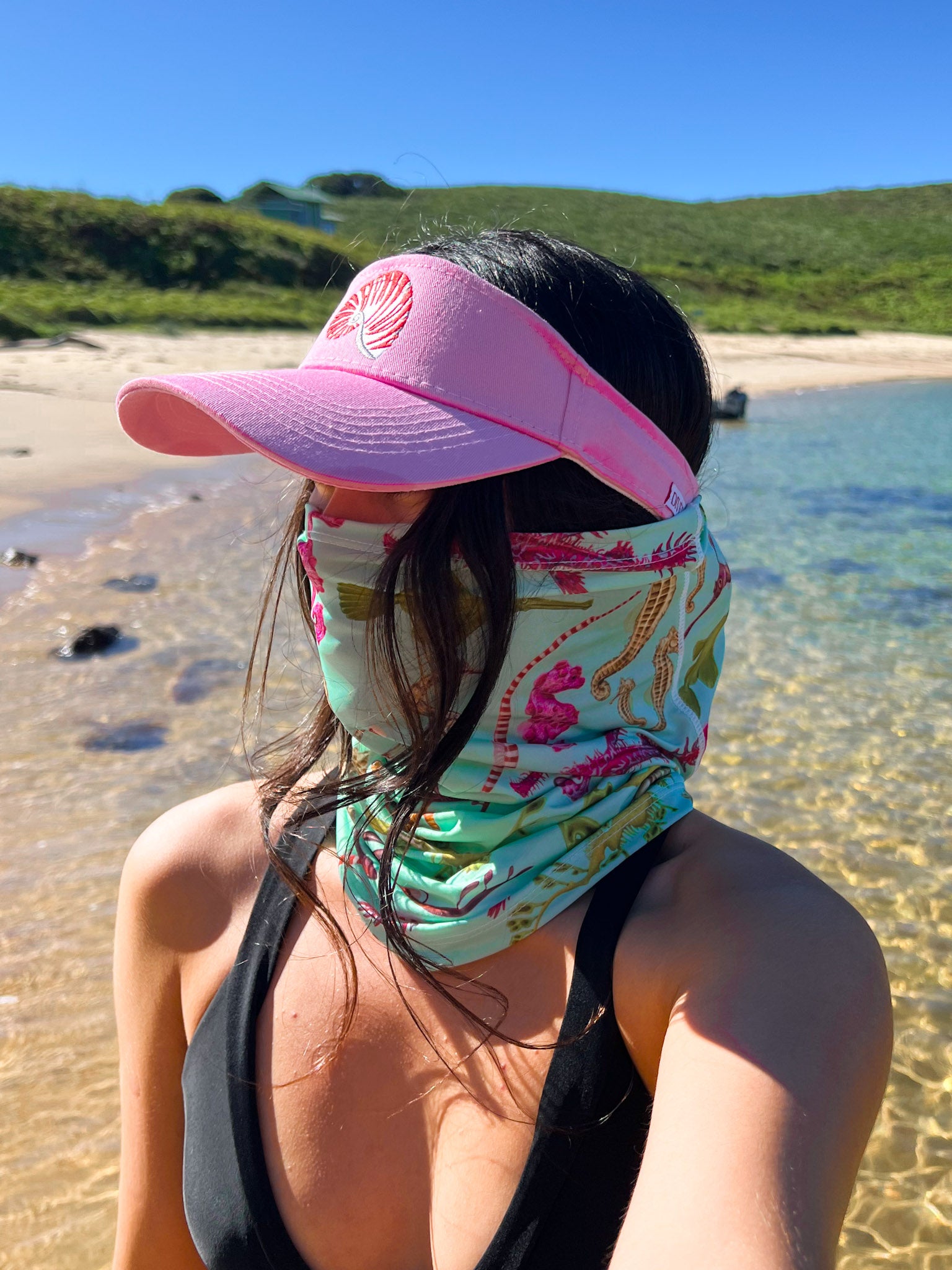 Dark haired women shown at the beach by the clear waters wearing the seahorse green UV gaiter buff made from recycled polyester-spandex around bottom half of face neck along with the pink nautilus visor hat ocean illustration by Diver Mon Art