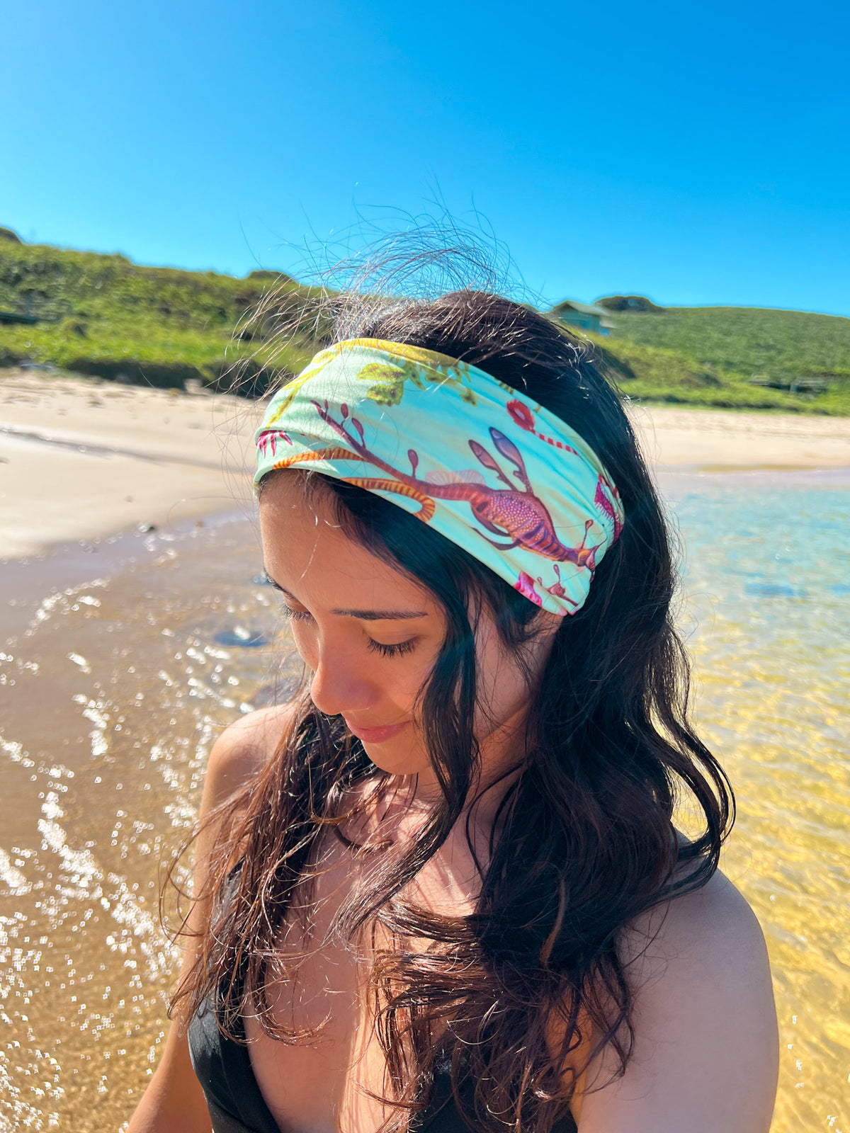 Dark haired women shown at the beach by the clear waters wearing the seahorse green UV gaiter buff made from recycled polyester-spandex headband style illustration by Diver Mon Art