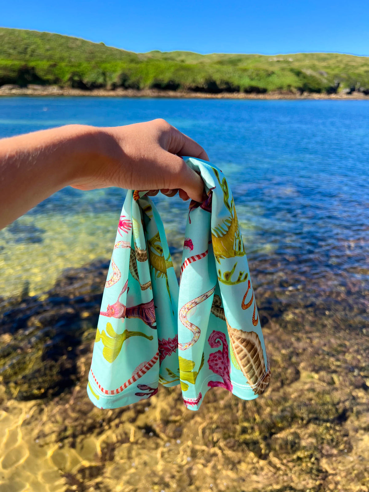 Seahorse green UV gaiter buff design shown held out by hand by the clear waters edge close up in great detail made from recycled polyester-spandex ocean illustration by Diver Mon Art