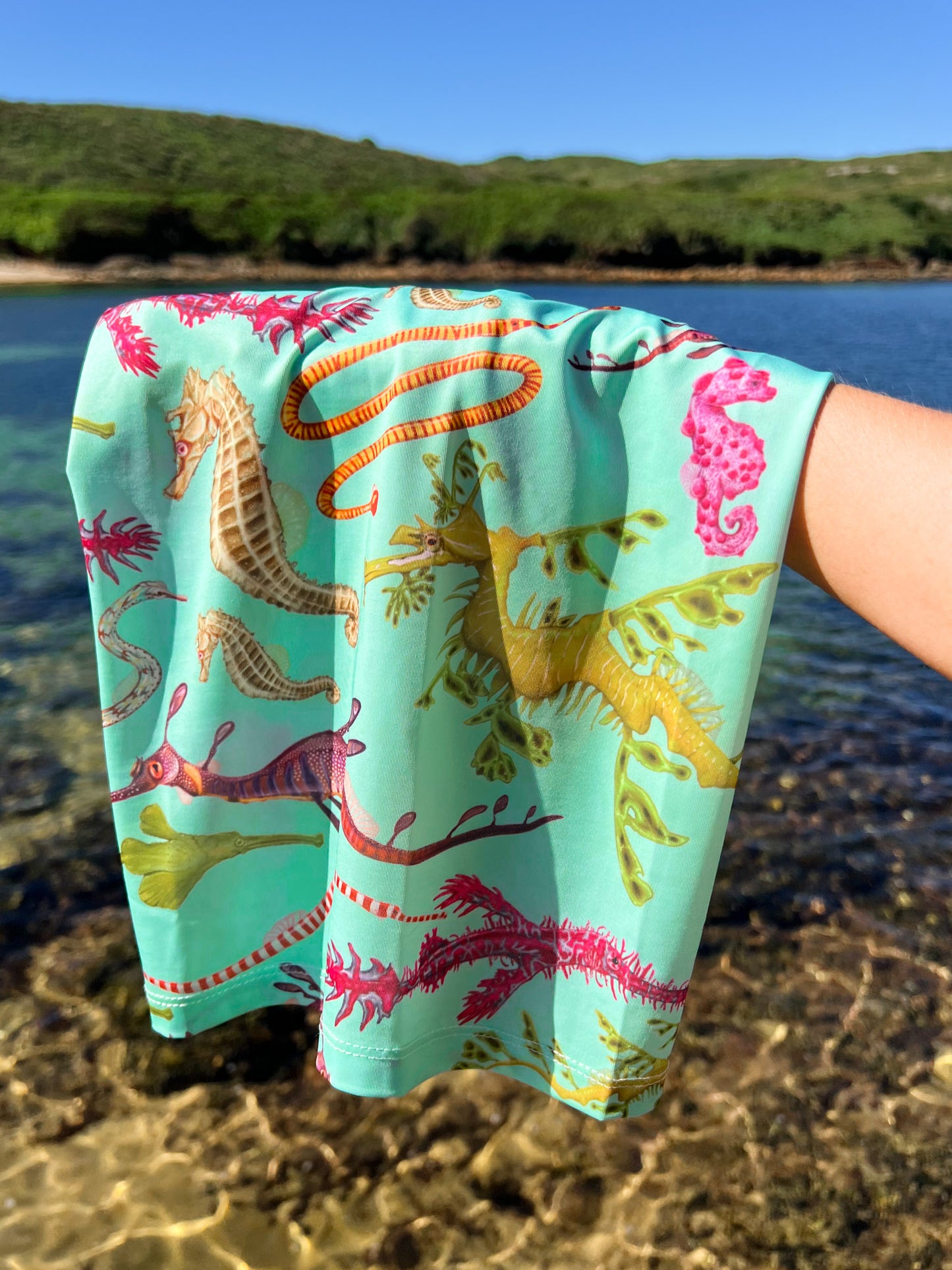 Seahorse green UV gaiter buff design shown hanging over hand by the clear waters edge close up in great detail made from recycled polyester-spandex ocean illustration by Diver Mon Art