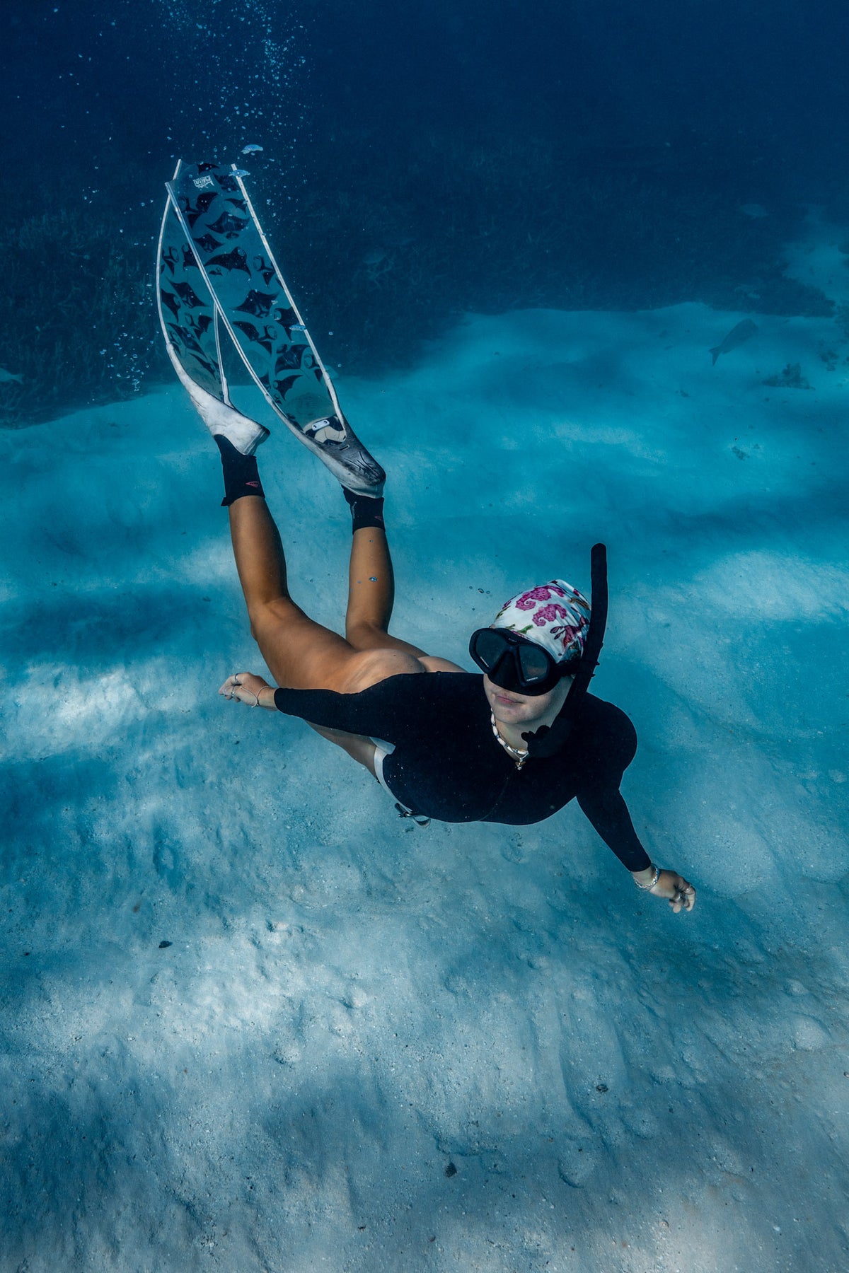 Blonde haired women shown from afar underwater clear blue water with manta white fins wearing seahorse green UV gaiter buff design made from recycled polyester-spandex ocean illustration by Diver Mon Art