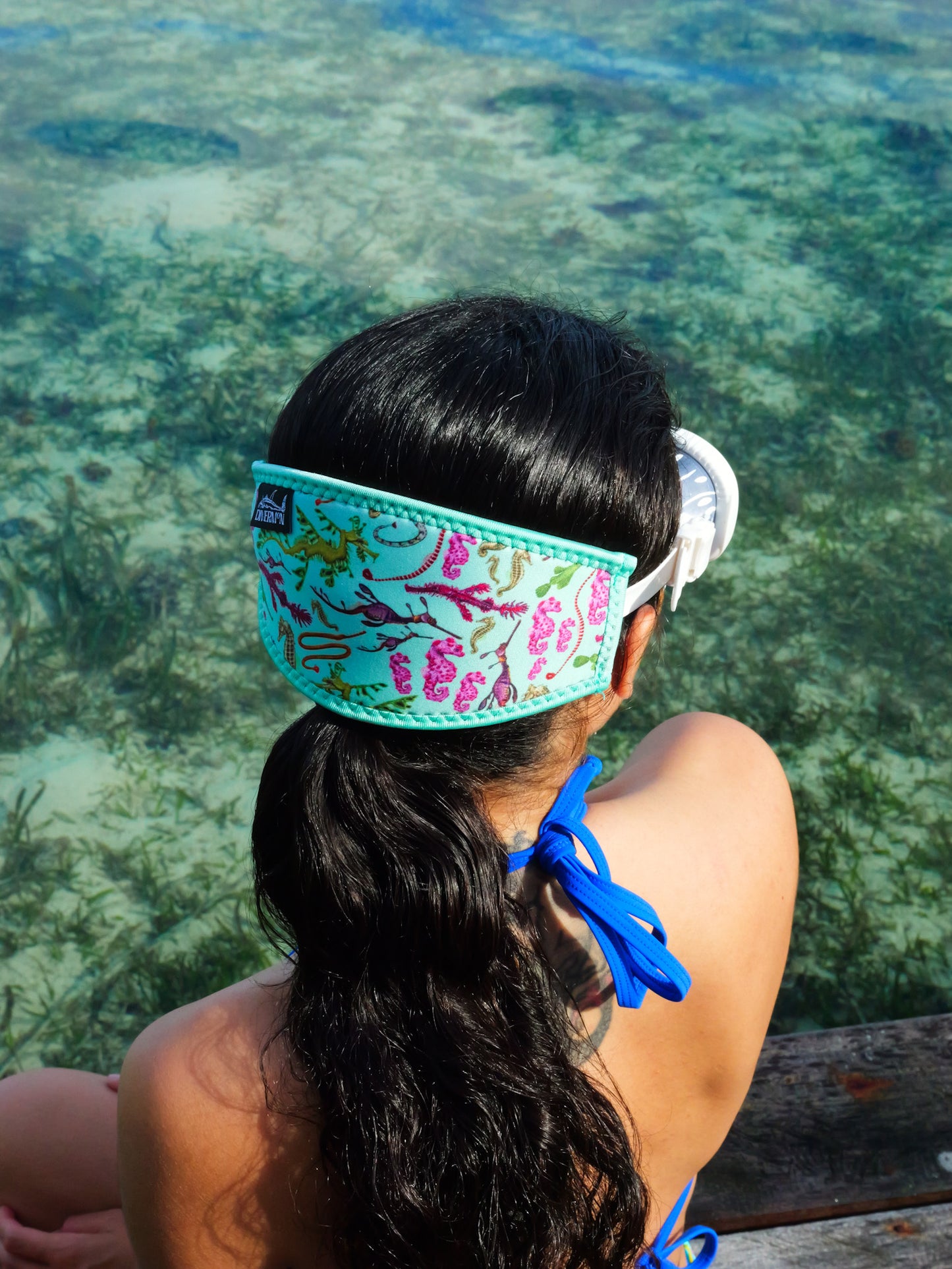 Dark haired woman shown from behind in blue clear water close up seahorse green design neoprene mask strap tamer fitted to mask ocean illustration by Diver Mon Art